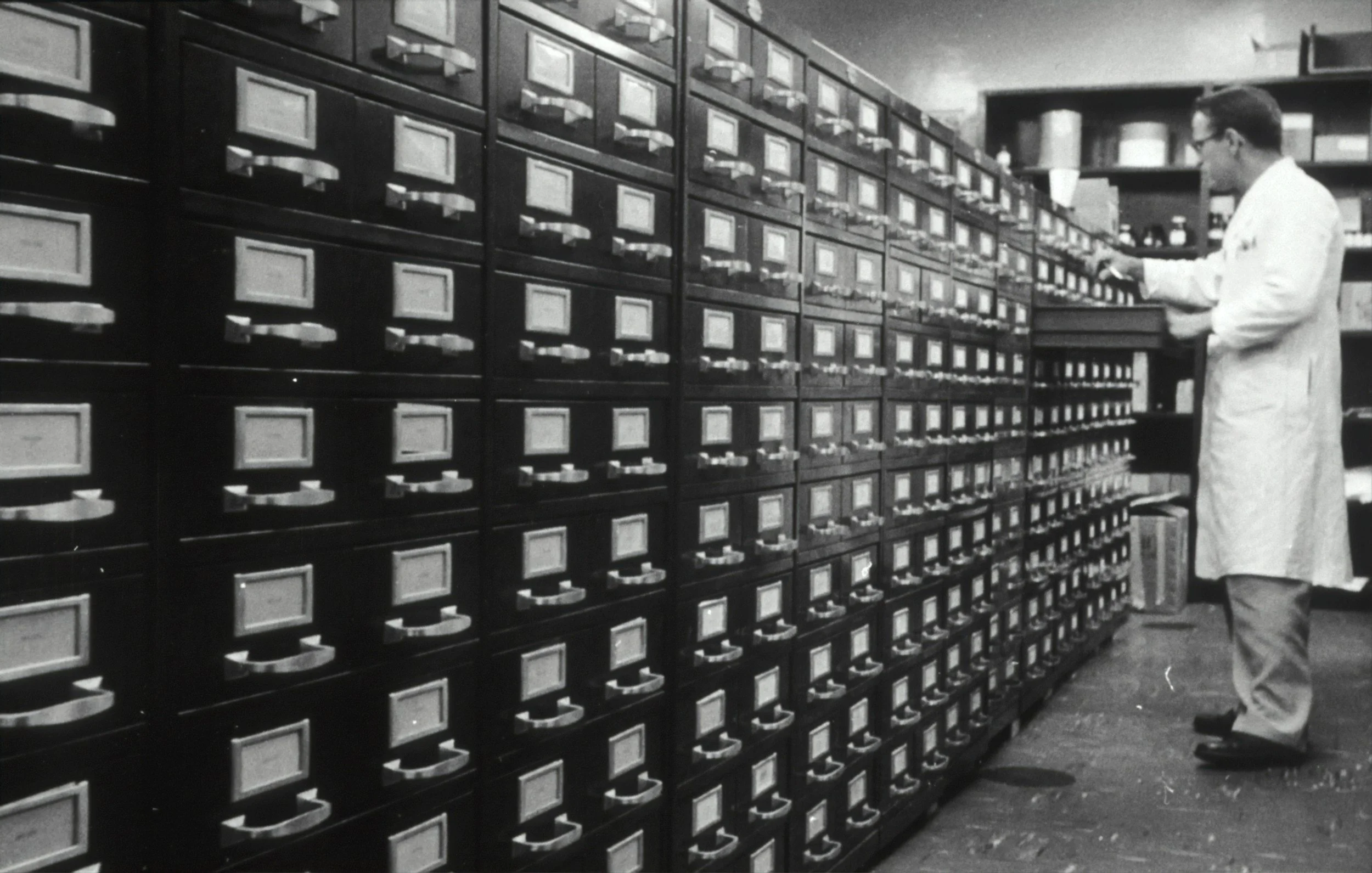 A man in a white lab coat working in a room with many filing cabinets or storage drawers.