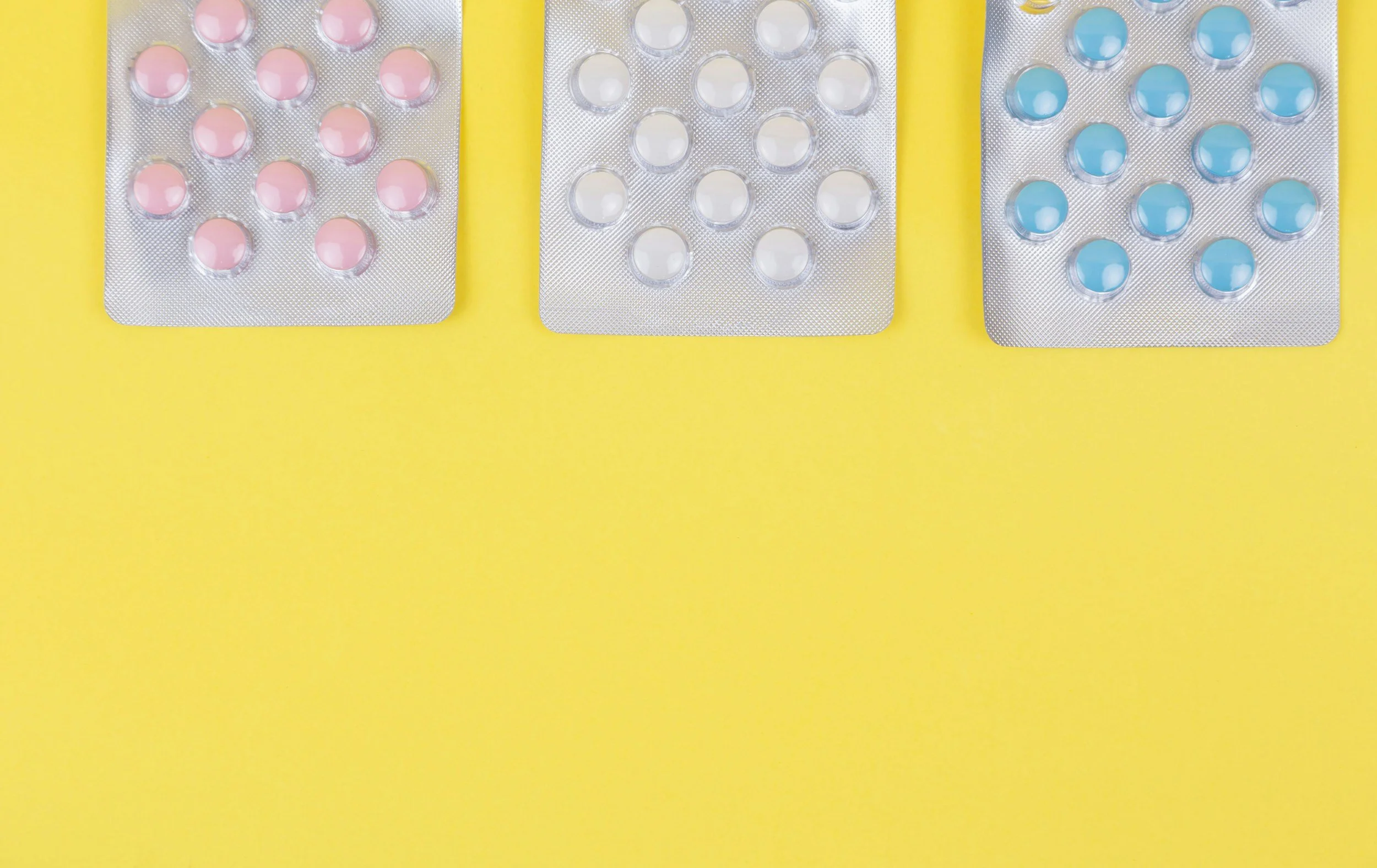 Three blister packs of pills, with pink, white, and blue pills, arranged on a yellow background.