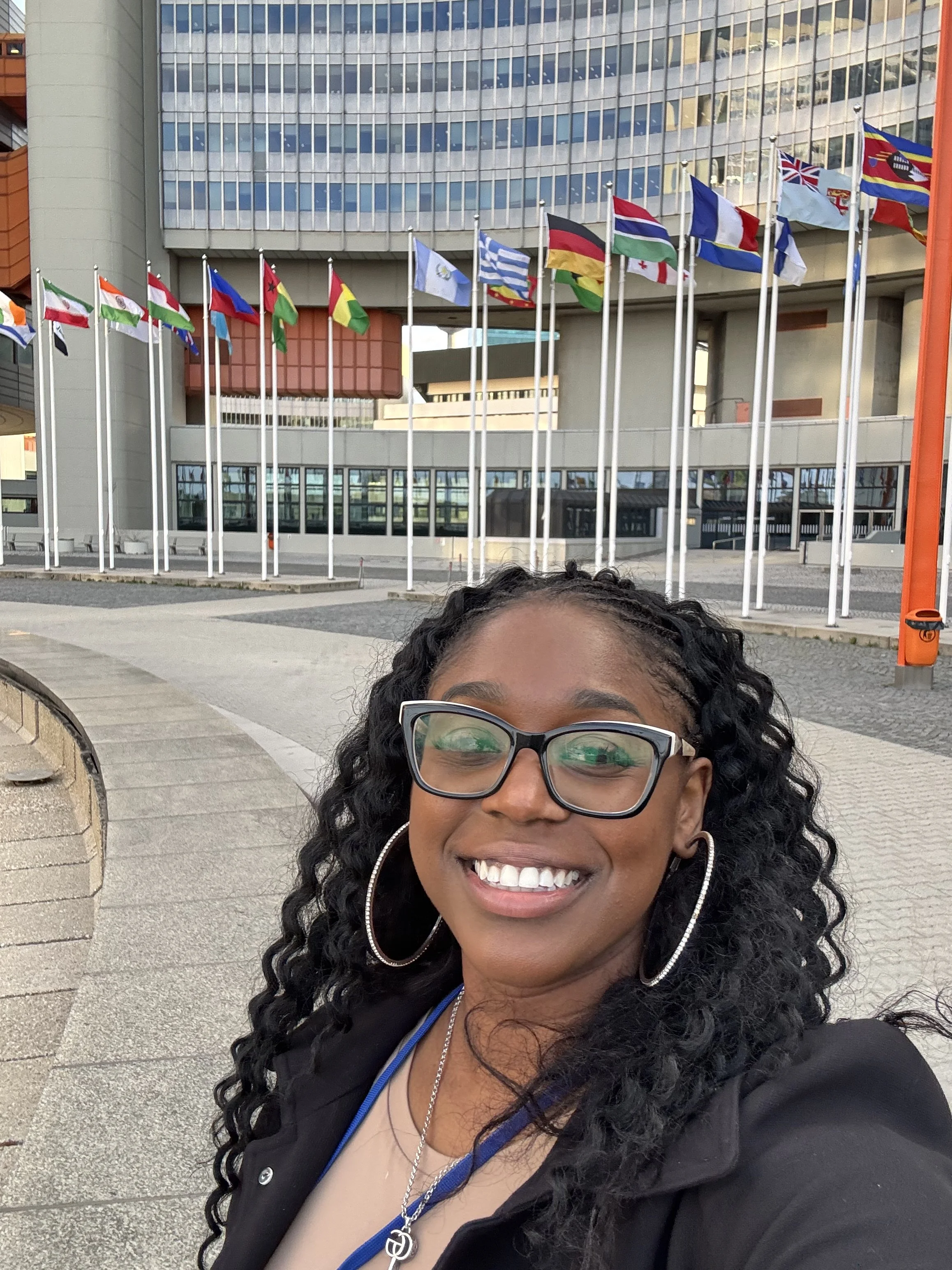 Smiling woman with glasses and hoop earrings taking a selfie in front of a building with multiple international flags.
