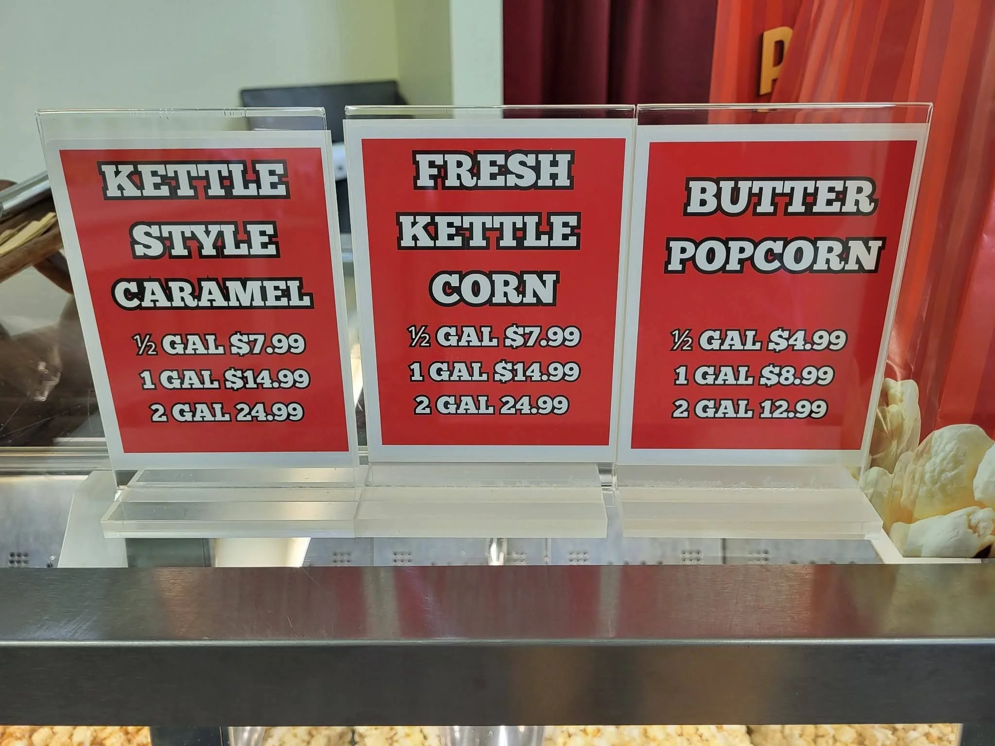 Red menu signs displaying prices for kettle style caramel, fresh kettle corn, and butter popcorn in a concession stand.