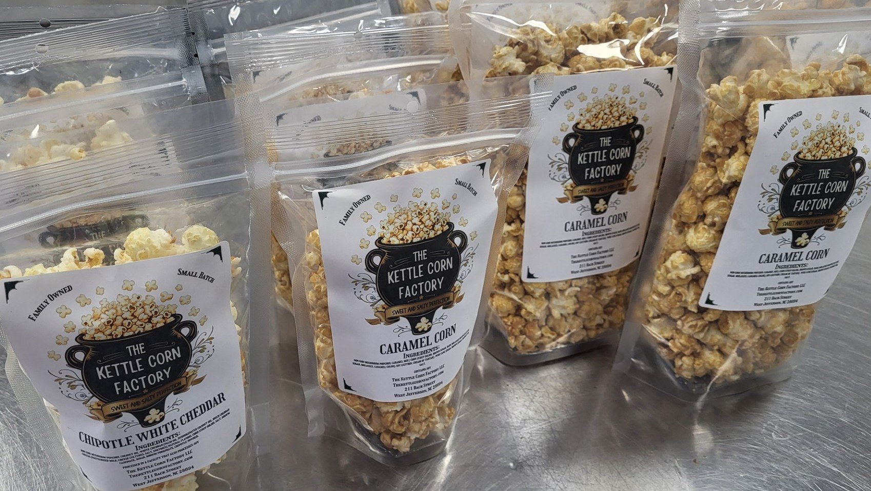 Several clear resealable bags of caramel popcorn with labels from The Kettle Corn Factory, featuring flavors Chipotle White Cheddar and Caramel Corn, placed on a metallic surface.