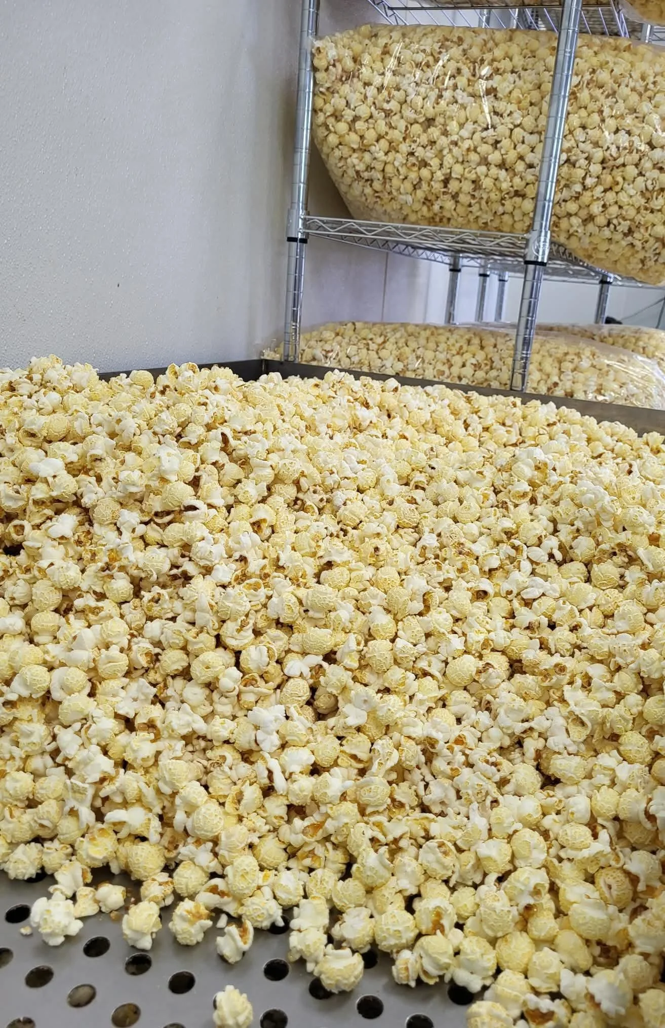 A large container of freshly popped popcorn on a perforated tray, with additional popcorn in clear plastic bags on a metal shelving unit behind.