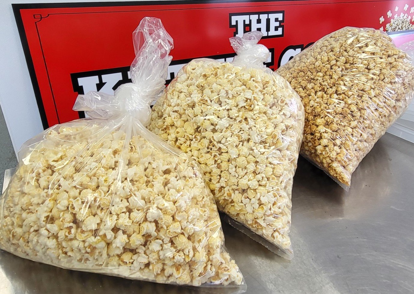Three large clear plastic bags filled with popcorn placed on a metallic surface, with a red and black sign in the background.