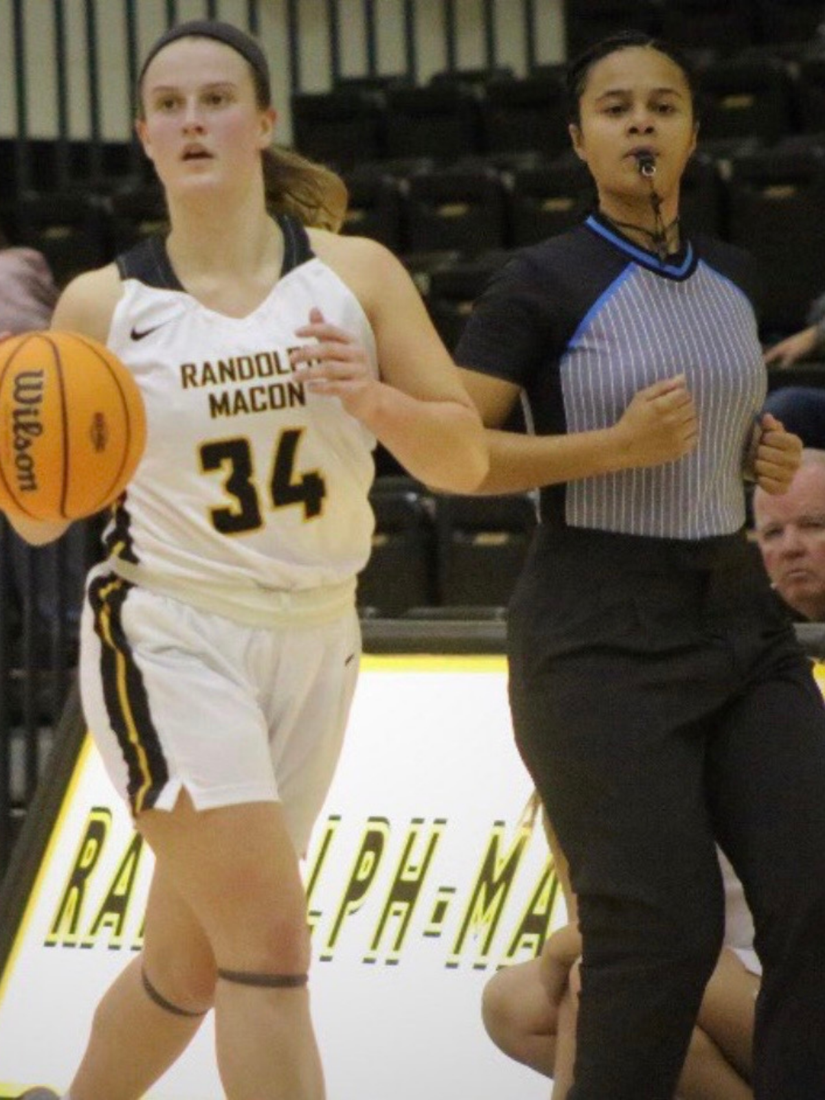 A female basketball player in a white uniform with 'Randolph Macon' and number 34, running on the court during a game, with a referee nearby.