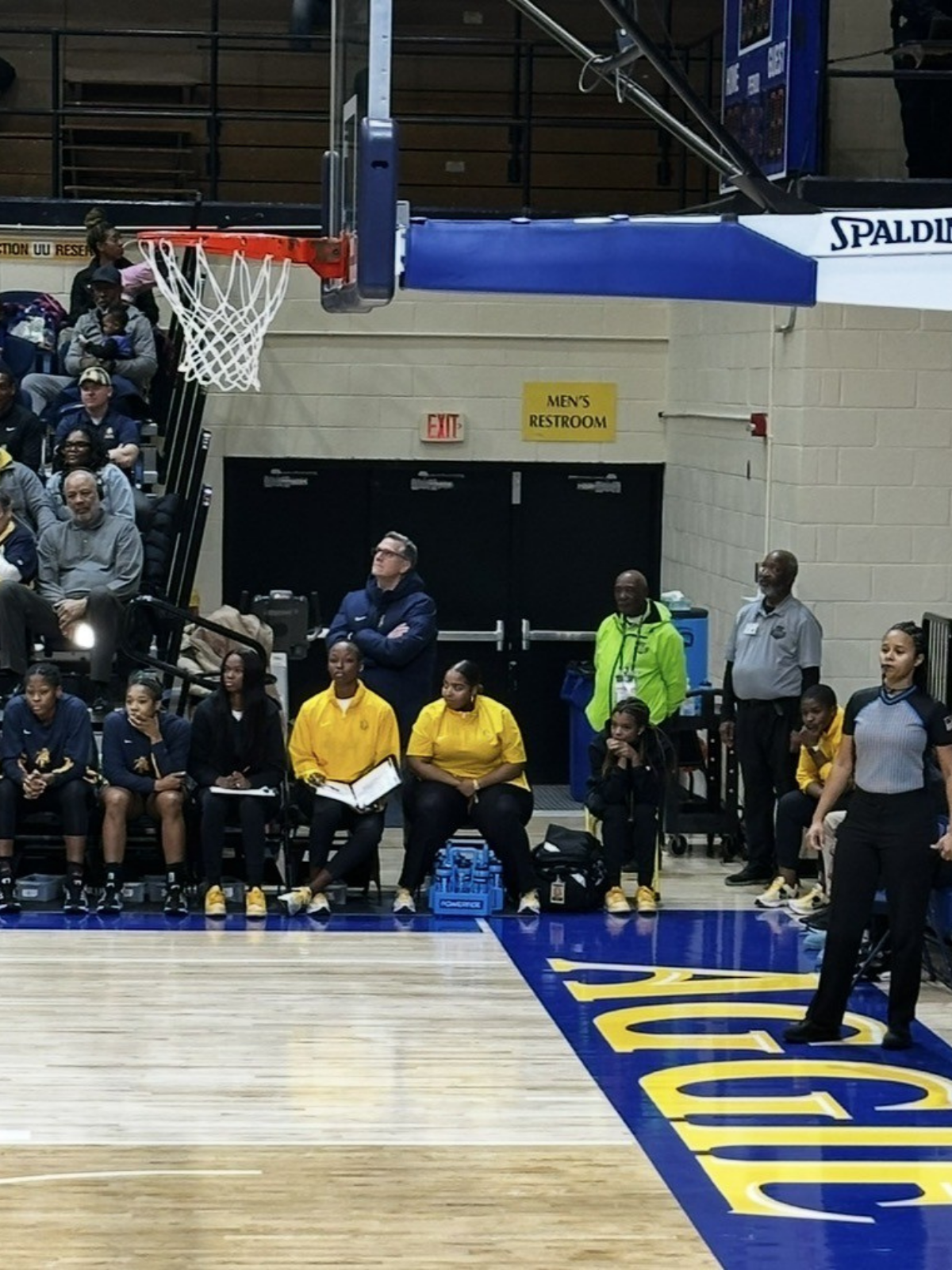 View of a basketball court with players sitting on the bench, coaches, and officials near the sideline. The scene appears to be during a game or event, with spectators in the background. The court has blue and yellow markings, and there's a visible section of a basketball hoop and backboard.