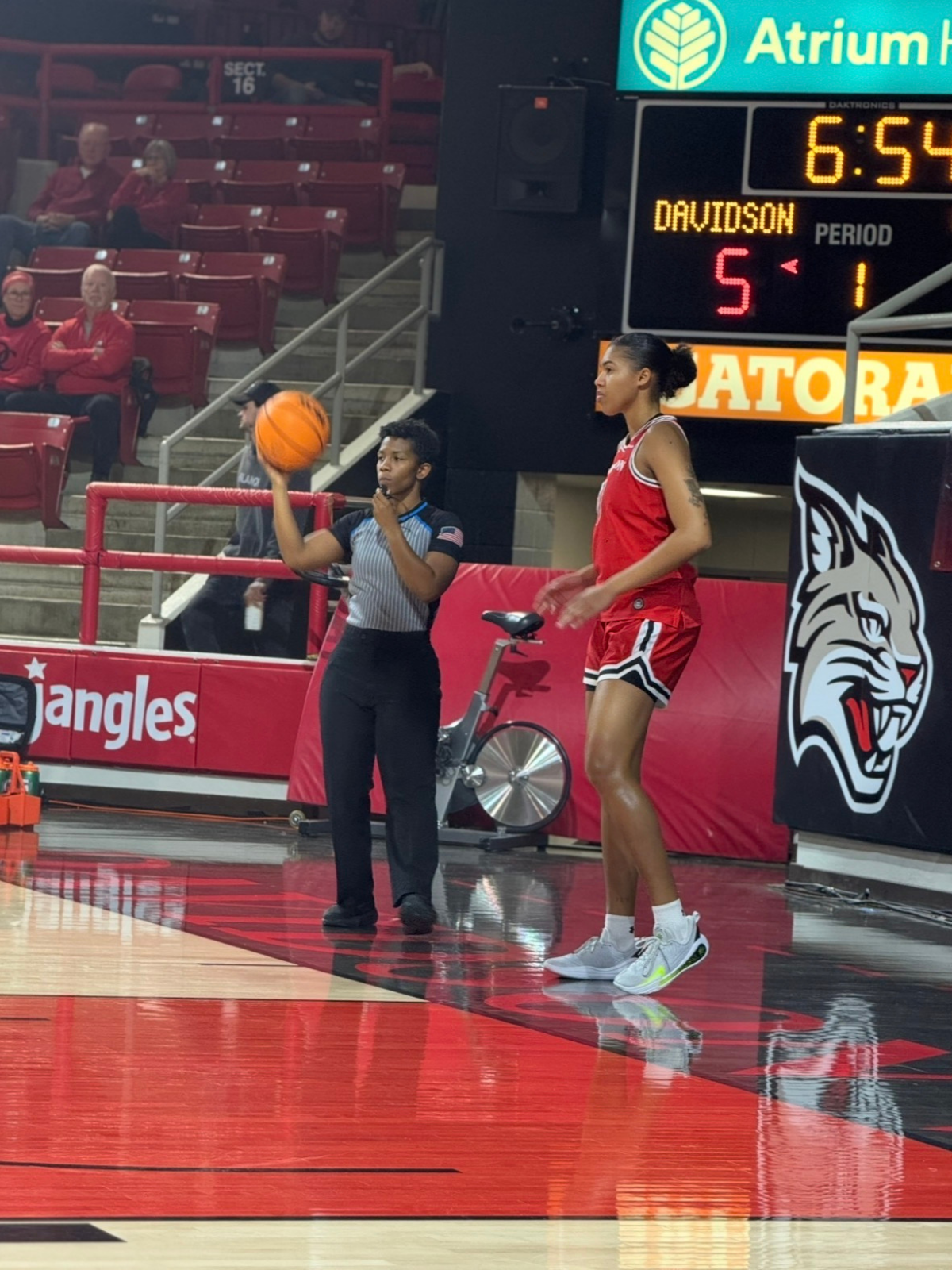 A female basketball player in a red uniform preparing to take a free throw on the basketball court, with a referee standing beside her holding a basketball, while spectators sit and watch in the background.