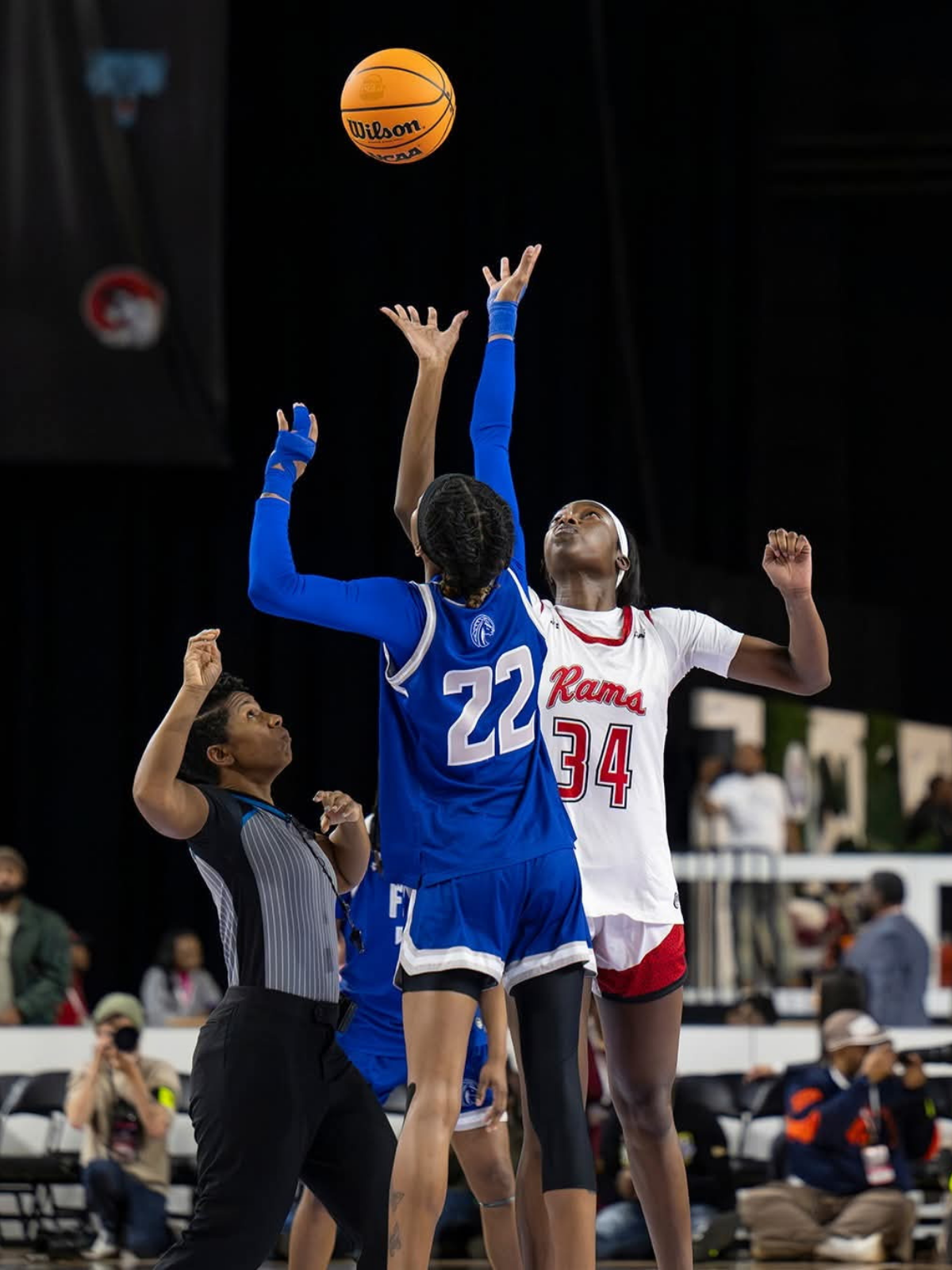 Two female basketball players, one in a blue uniform and the other in a white and red uniform, jump to reach a basketball in mid-air during a game, with a referee watching nearby.