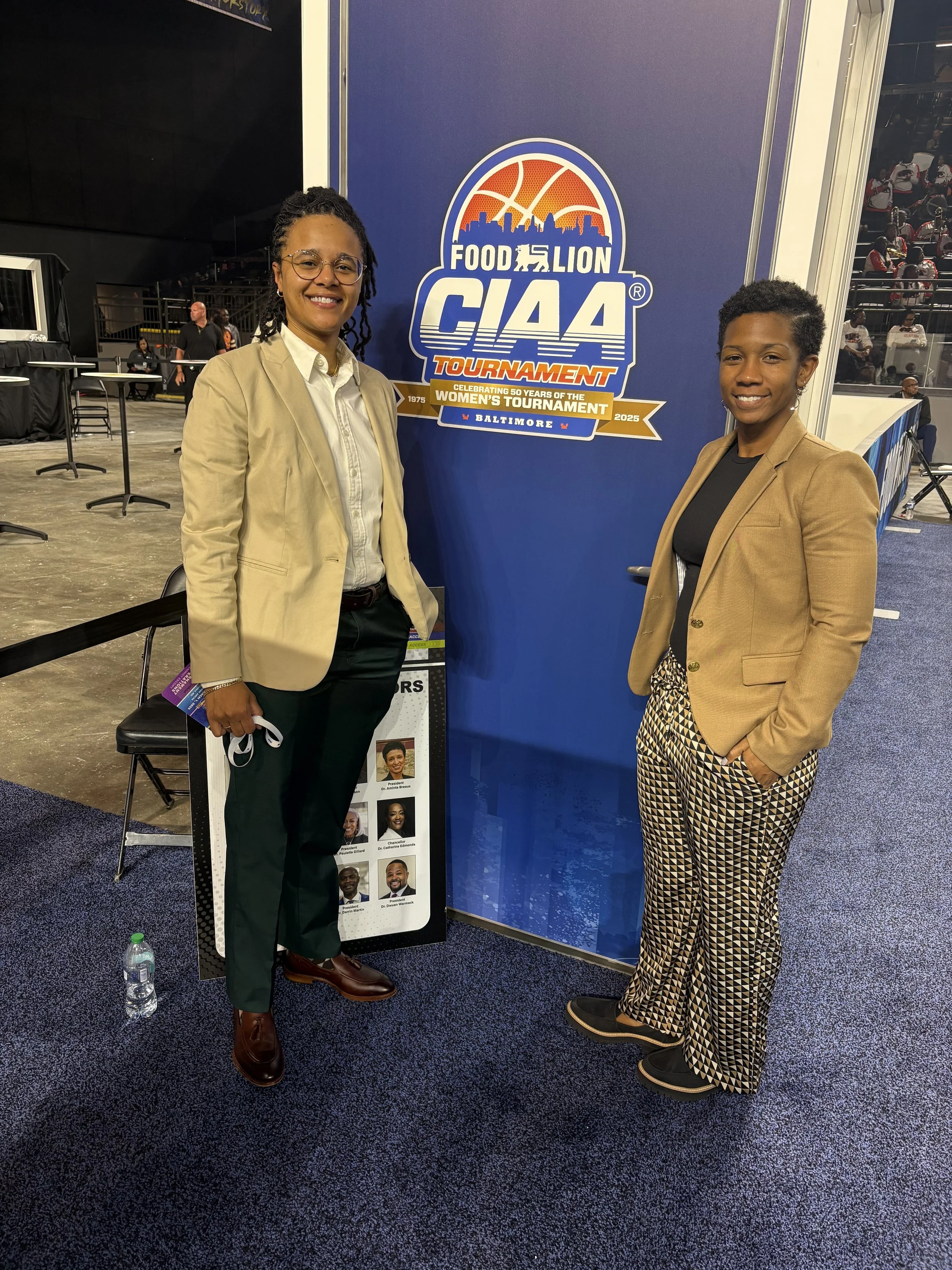 Two women standing indoors, smiling, posing for a photo in front of a large blue sign that reads 'Food Lion CIAA Tournament 2025 Baltimore'. The woman on the left is holding a face mask and a water bottle on the floor next to her, and the woman on the right has her hand in her pocket. There is a display with photos and names of people beneath the sign. In the background, there are chairs and a crowd of people.