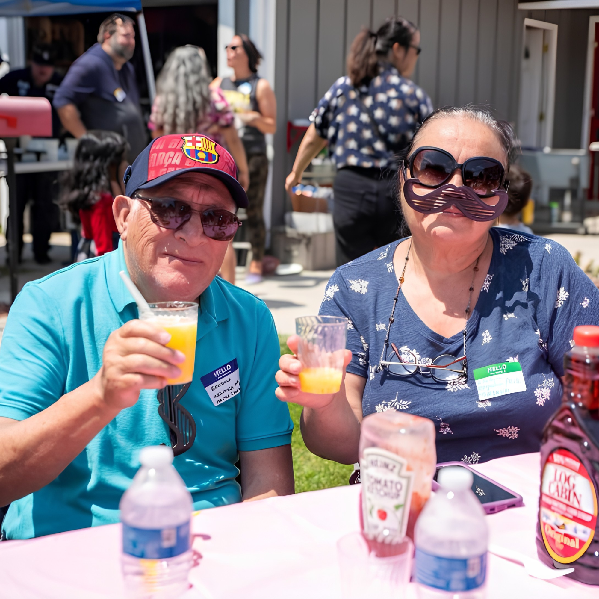 Two older adults, a man and a woman, sitting at a table outdoors at a social gathering, clinking glasses with orange drinks and wearing funny sunglasses with mustache props, smiling.