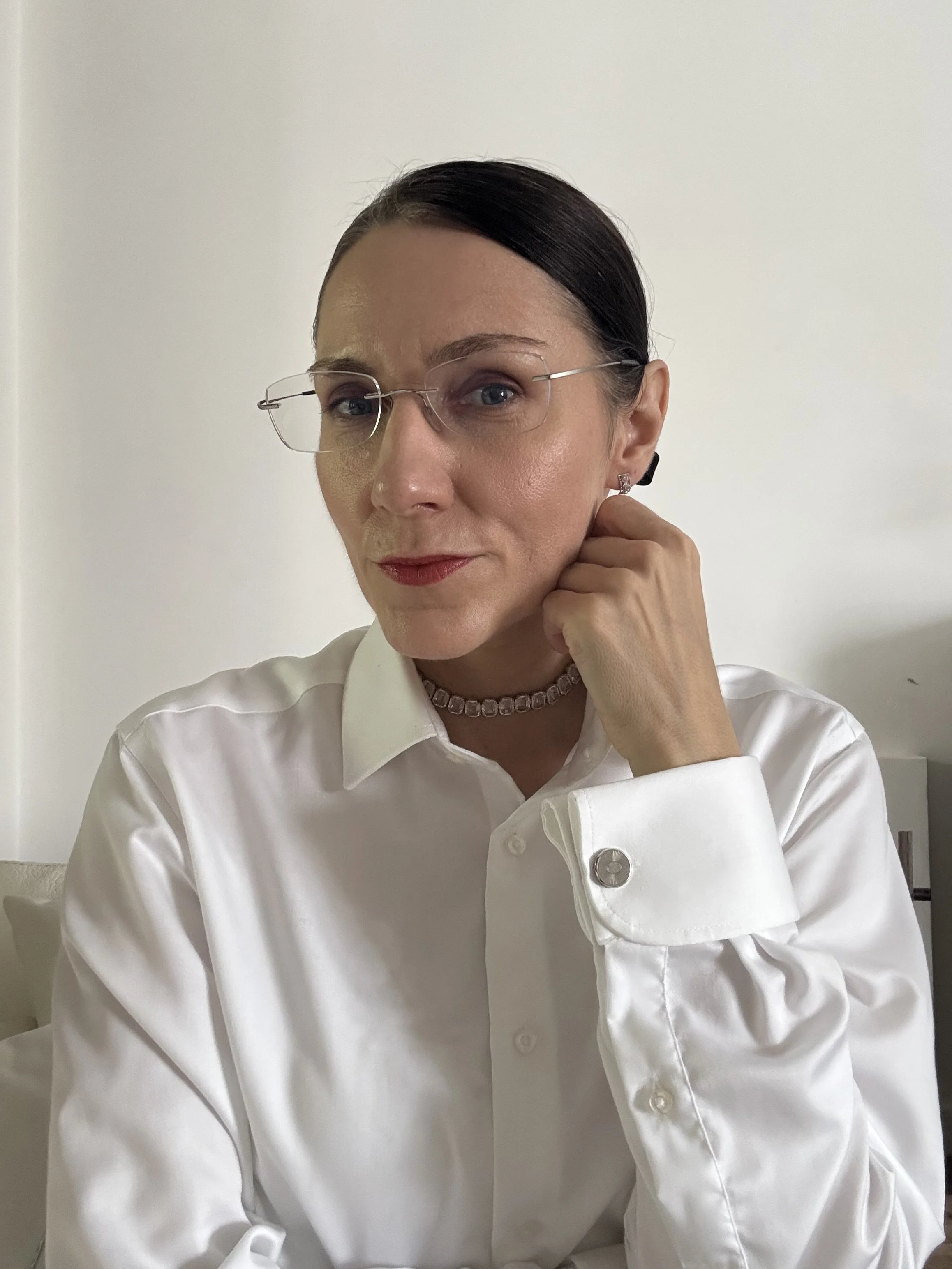 A woman with dark hair pulled back, wearing glasses, a white button-up shirt, a choker necklace, and earrings, sitting with her hand resting on her jaw against a plain white background.