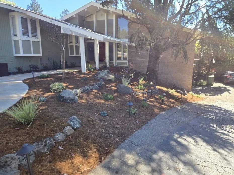 A landscaped front yard with mulch, rocks, and small plants, in front of a house with large windows and a covered porch. There is a tree casting shadows on the driveway.