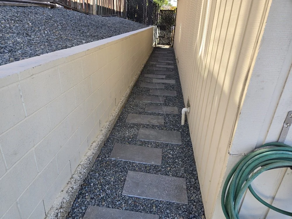 Narrow outdoor side pathway lined with a beige wall on the right and a white cinder block wall on the left, with stepping stones on gravel, leading to a gate.