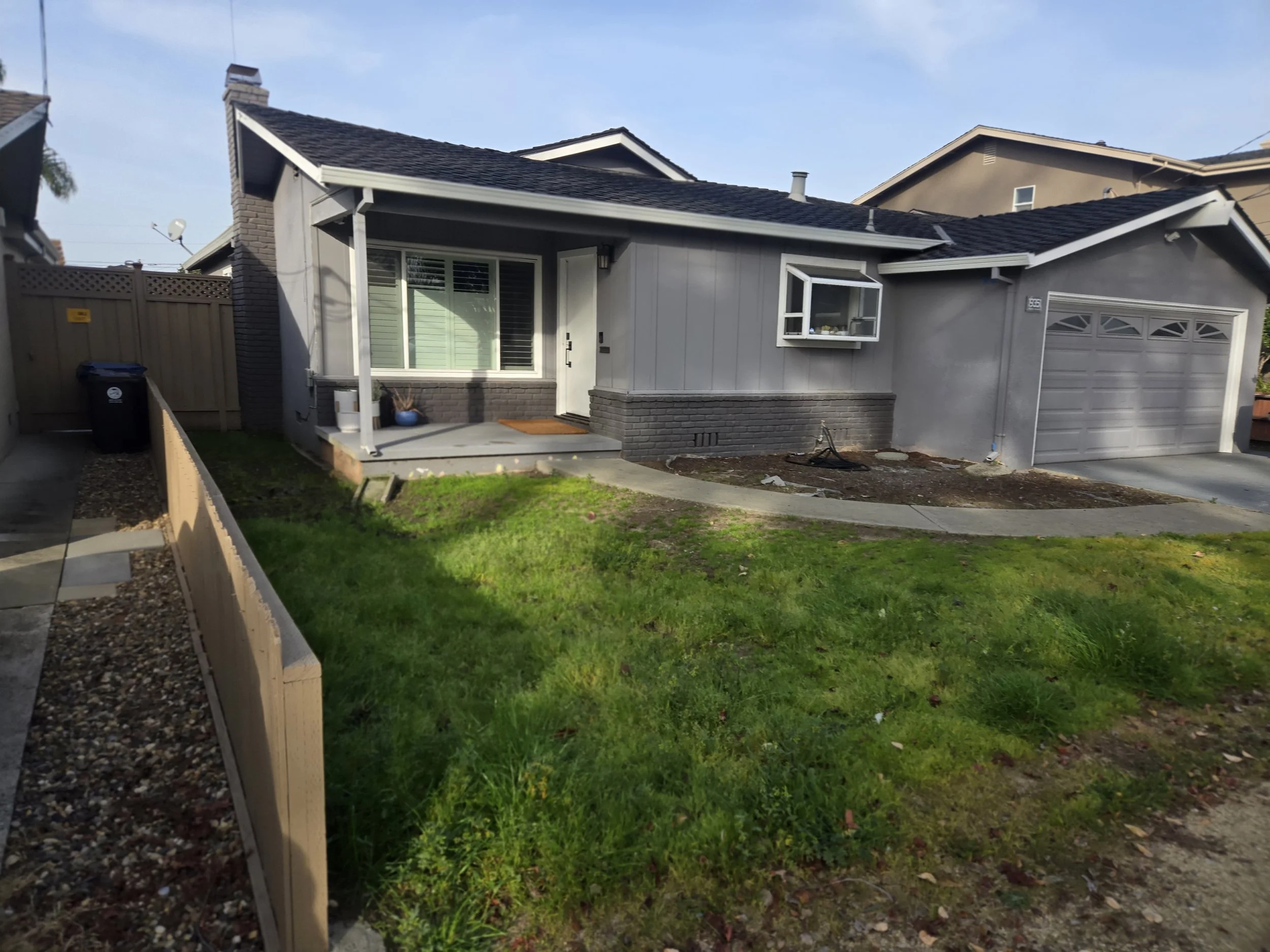 A suburban house with a small front yard, a porch, and a driveway, with neighboring houses visible in the background. The yard has a grass patch and a wooden fence on the left. The house has gray siding, a brick foundation, and a black-shingled roof.