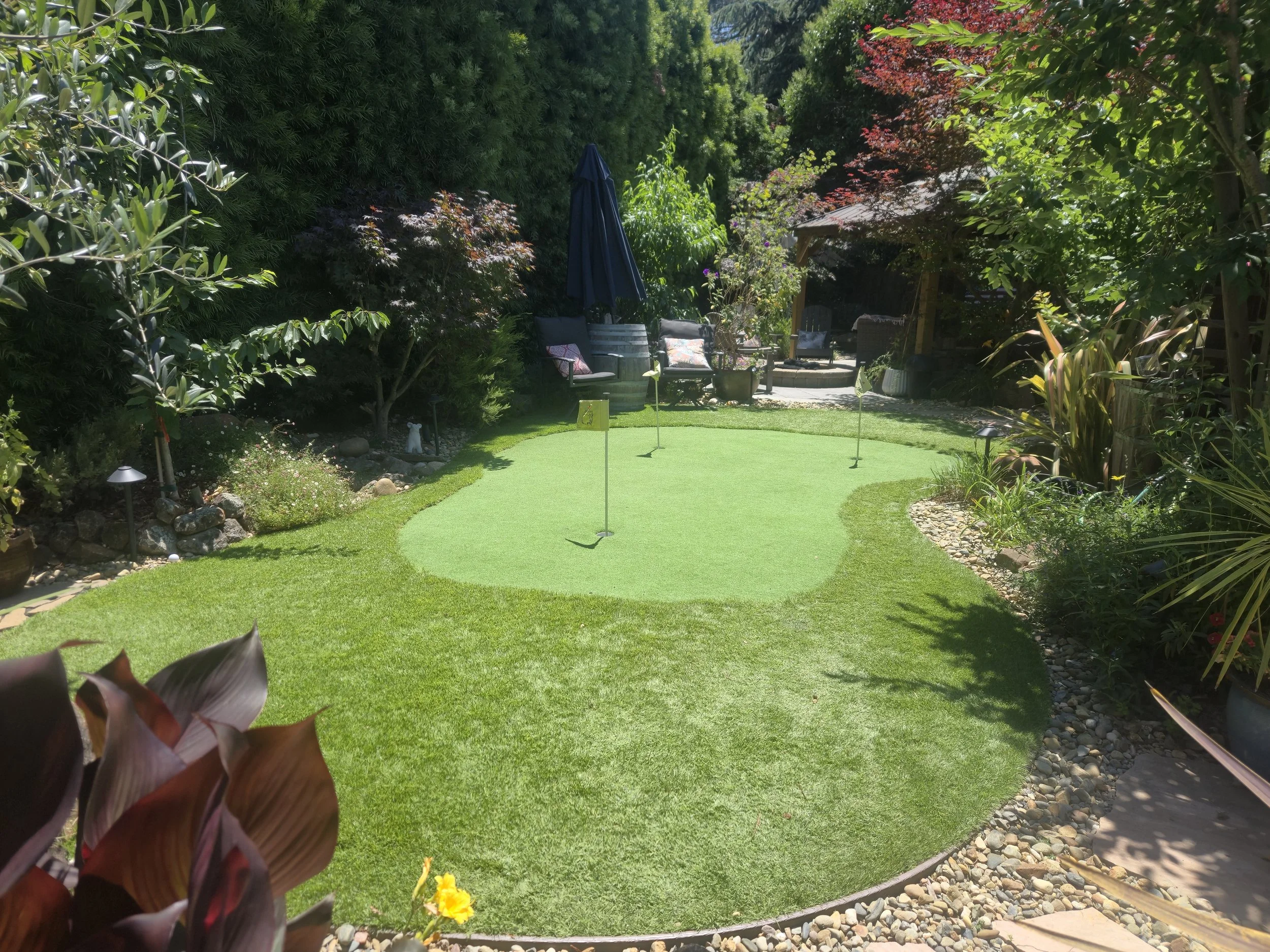 A backyard putting green with flags, surrounded by lush green trees and plants, along with outdoor seating and a shaded area.