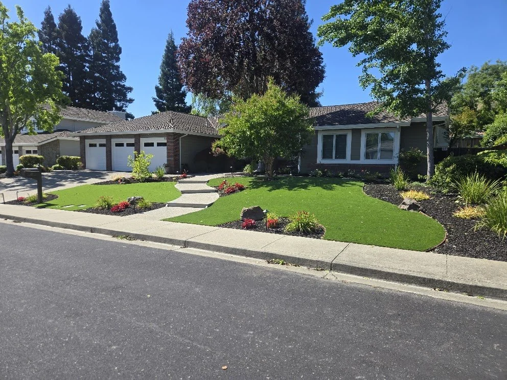 A suburban home with a well-maintained front yard that features lush green artificial grass, flower beds with red and pink flowers, and several trees. The house has a brick and siding exterior, a driveway with two garage doors, and a sidewalk along t