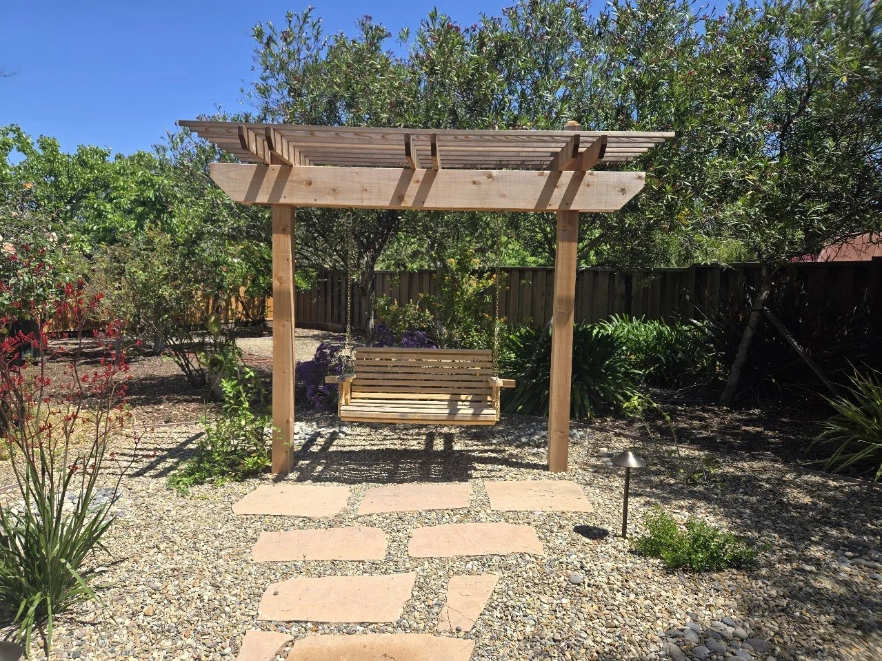 A backyard garden with a wooden swing with a roof, set on a gravel pathway with stepping stones, surrounded by green plants and trees, under a clear blue sky.