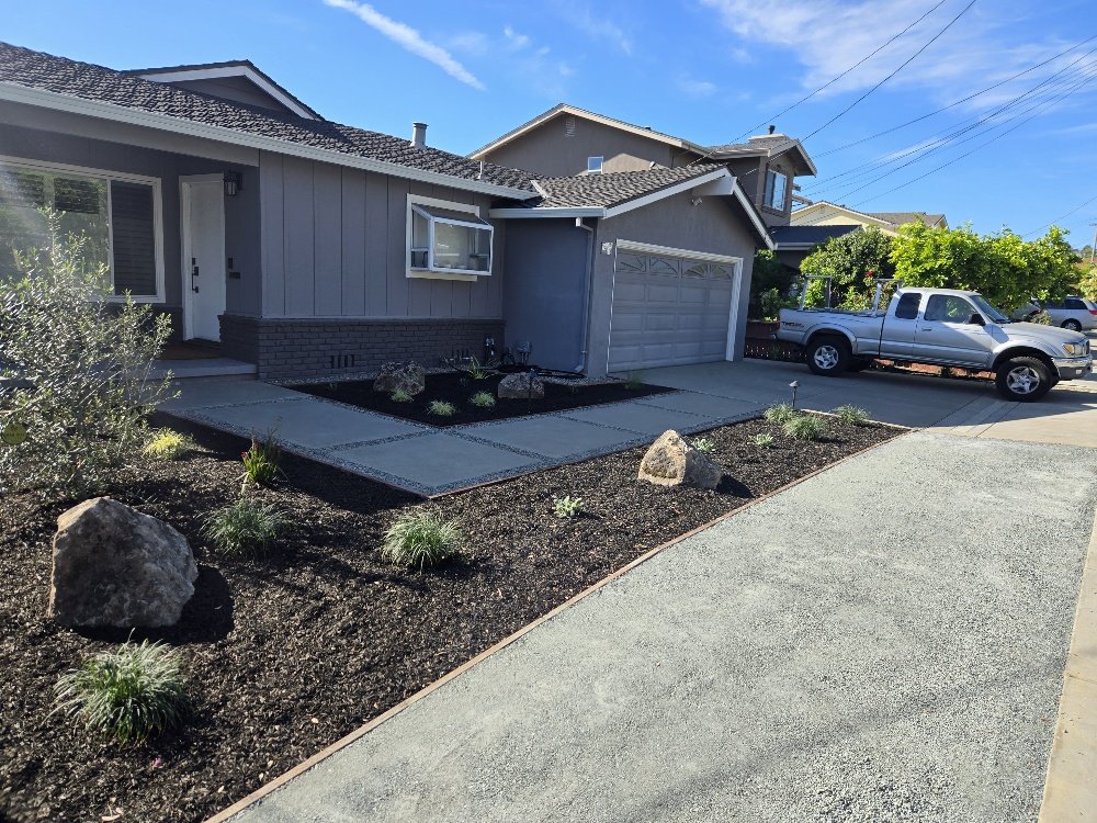 Front yard of a house with a small garden bed, rocks, and plants, concrete walkway, driveway, gray house with a garage, white pickup truck parked nearby, and a clear blue sky.