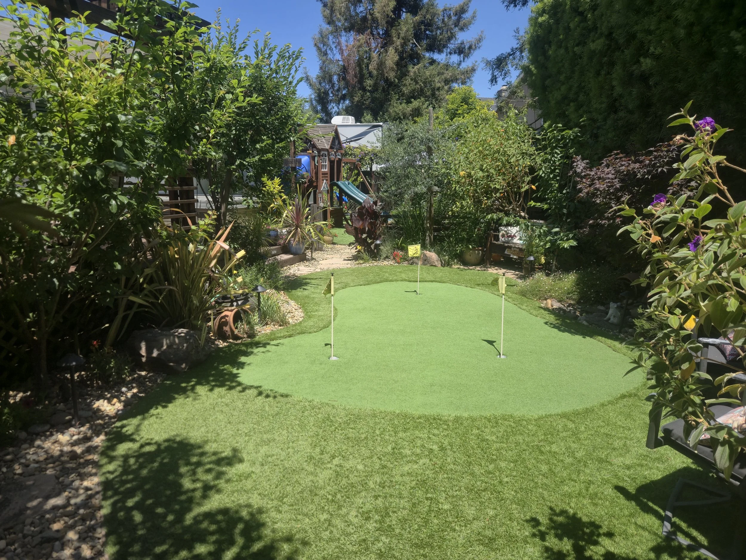 A backyard garden with a putting green, surrounded by lush greenery, trees, and flowering plants under a clear blue sky.