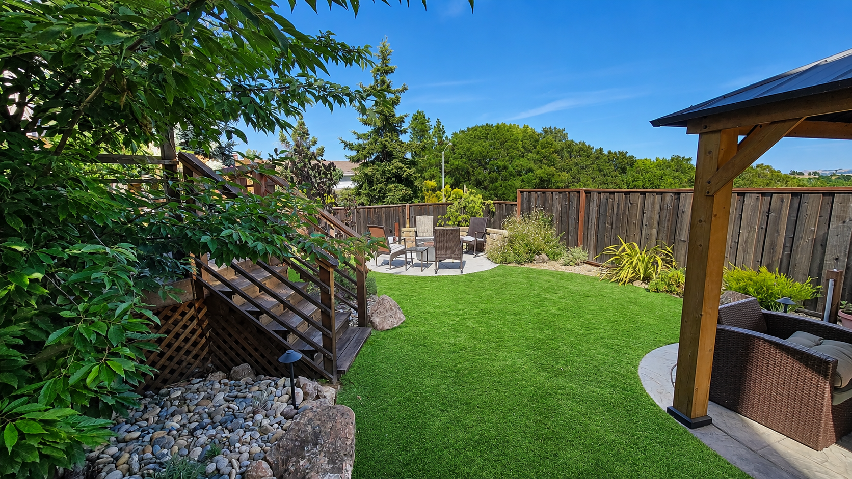 A backyard with a green lawn, patio furniture, a wooden staircase leading to a deck, a shaded seating area under a pergola, and a wooden fence all around, with trees and a blue sky in the background.