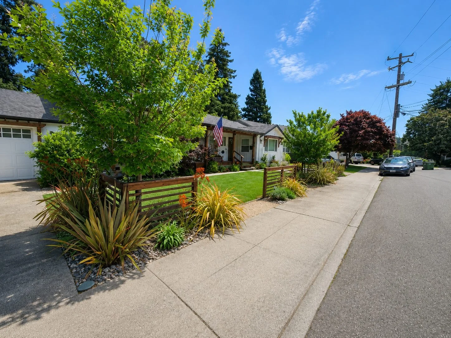 A suburban street with a single-story house, lush front yard with various trees and plants, a small fenced garden, parked cars, utility poles, and a clear blue sky.