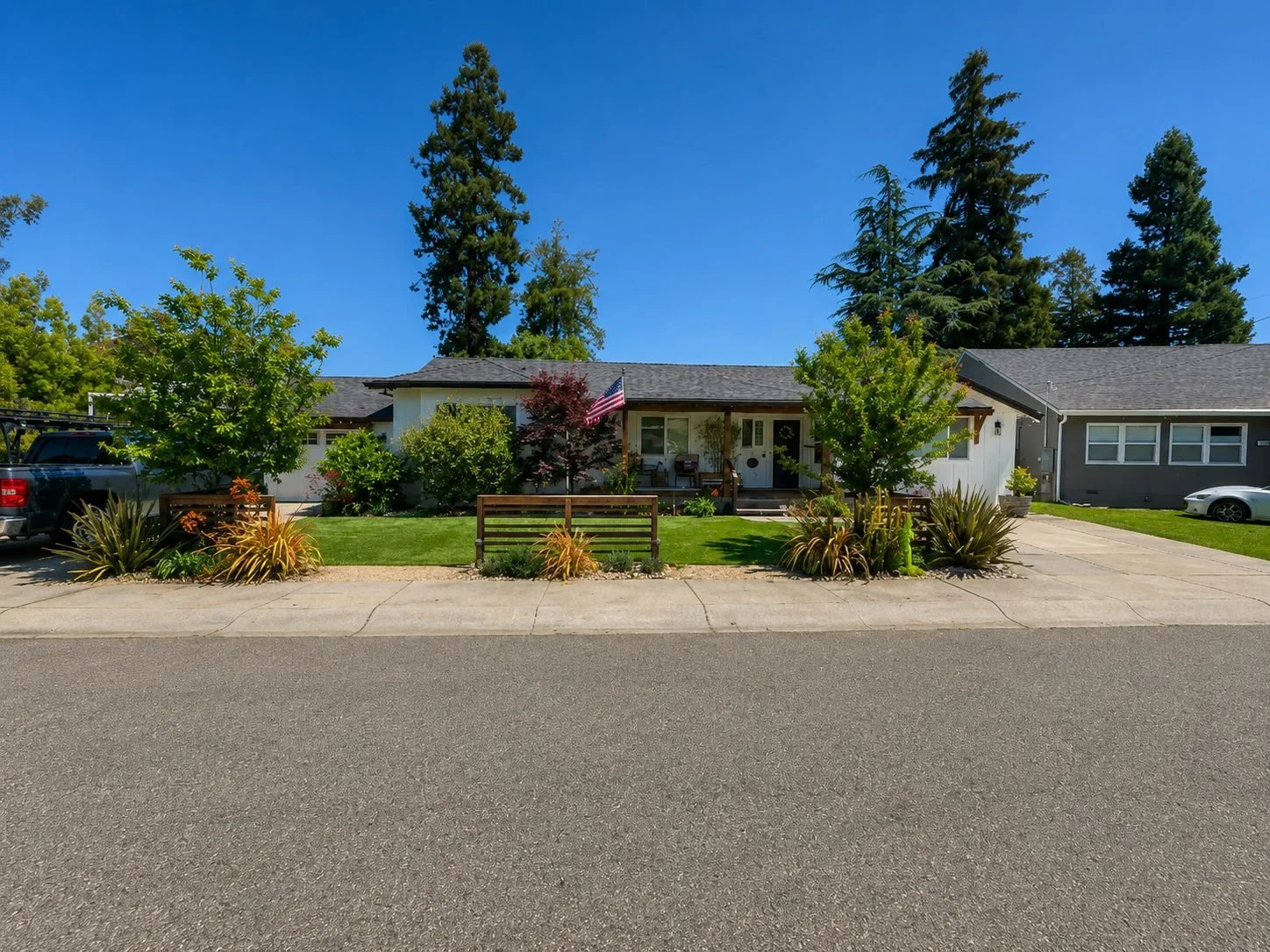 Front view of a single-story house with a well-maintained lawn, trees, and shrubs, with a driveway and parked vehicles on either side, under a clear blue sky.