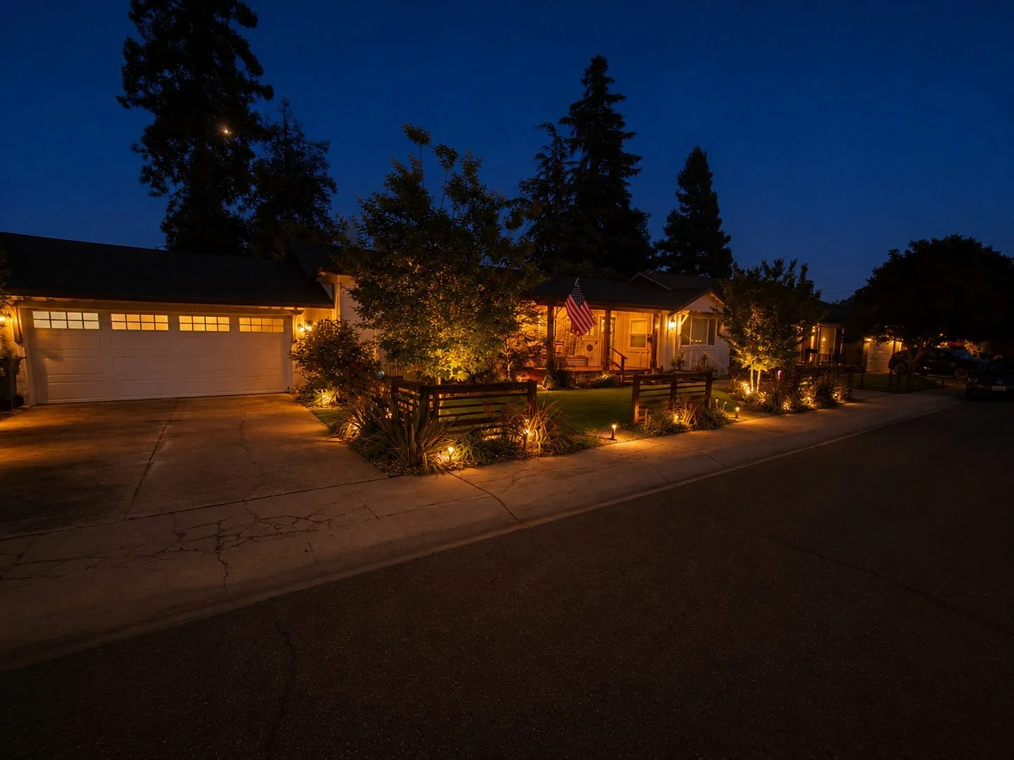 Night view of suburban houses with warm outdoor lighting, trees, and an American flag on the front porch.