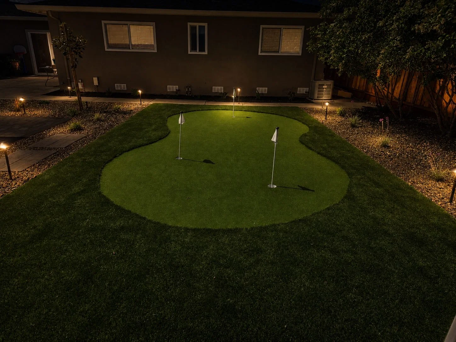 Nighttime backyard with a mini golf putting green, three small flags, and garden lights along the pathway