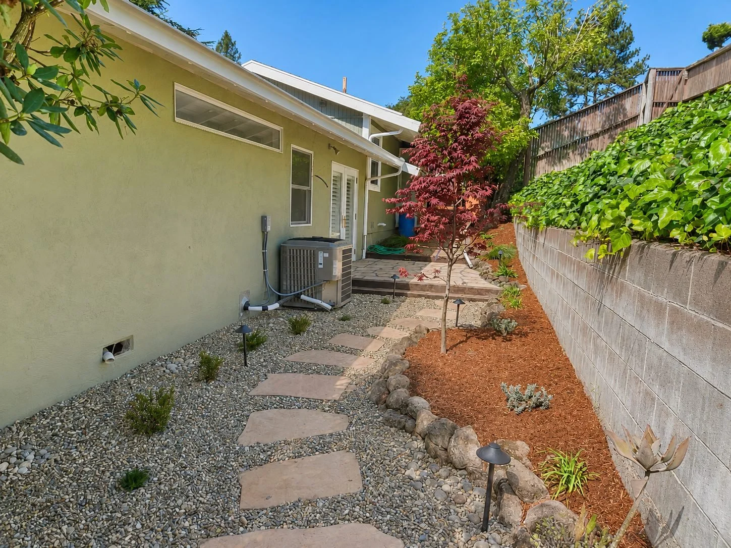 Backyard with stepping stones, small plants, trees, and a concrete wall, next to a house with a green exterior and an air conditioning unit.