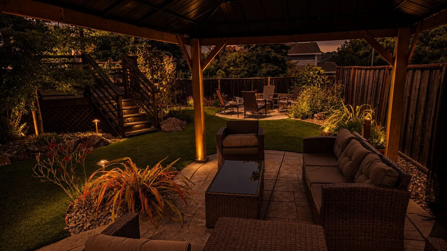 Illuminated backyard patio area at dusk with wicker furniture, a fire pit, a lawn, and a raised seating area, enclosed by a wooden fence and surrounded by trees and shrubs.