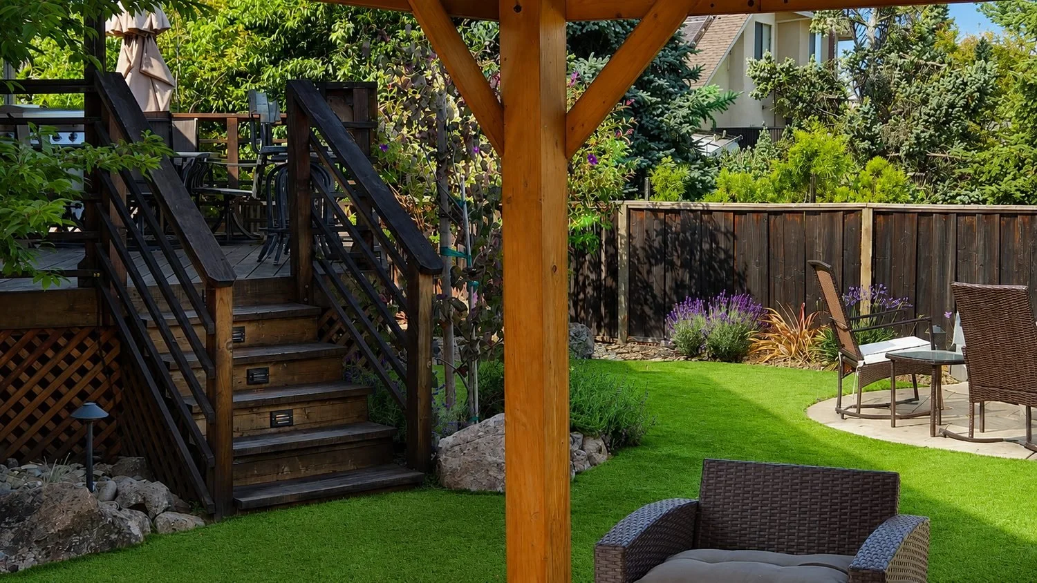 Backyard garden with patio, patio furniture, elevated wooden deck, lush green grass, trees, shrubs, and flowering plants.