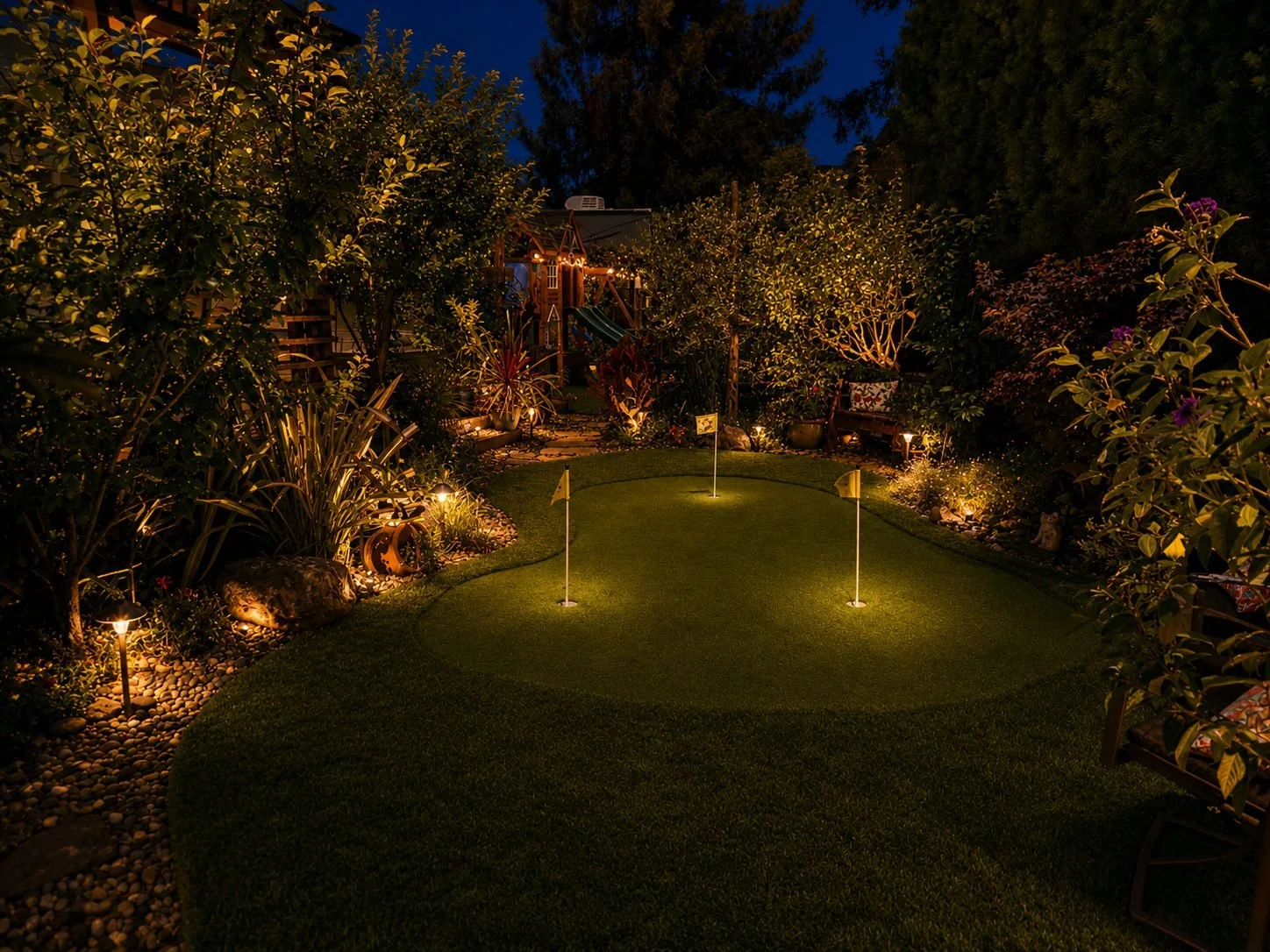 A backyard at night featuring a small putting green with three yellow flags, surrounded by decorative plants, shrubs, and garden lights, with a children's play structure in the background.