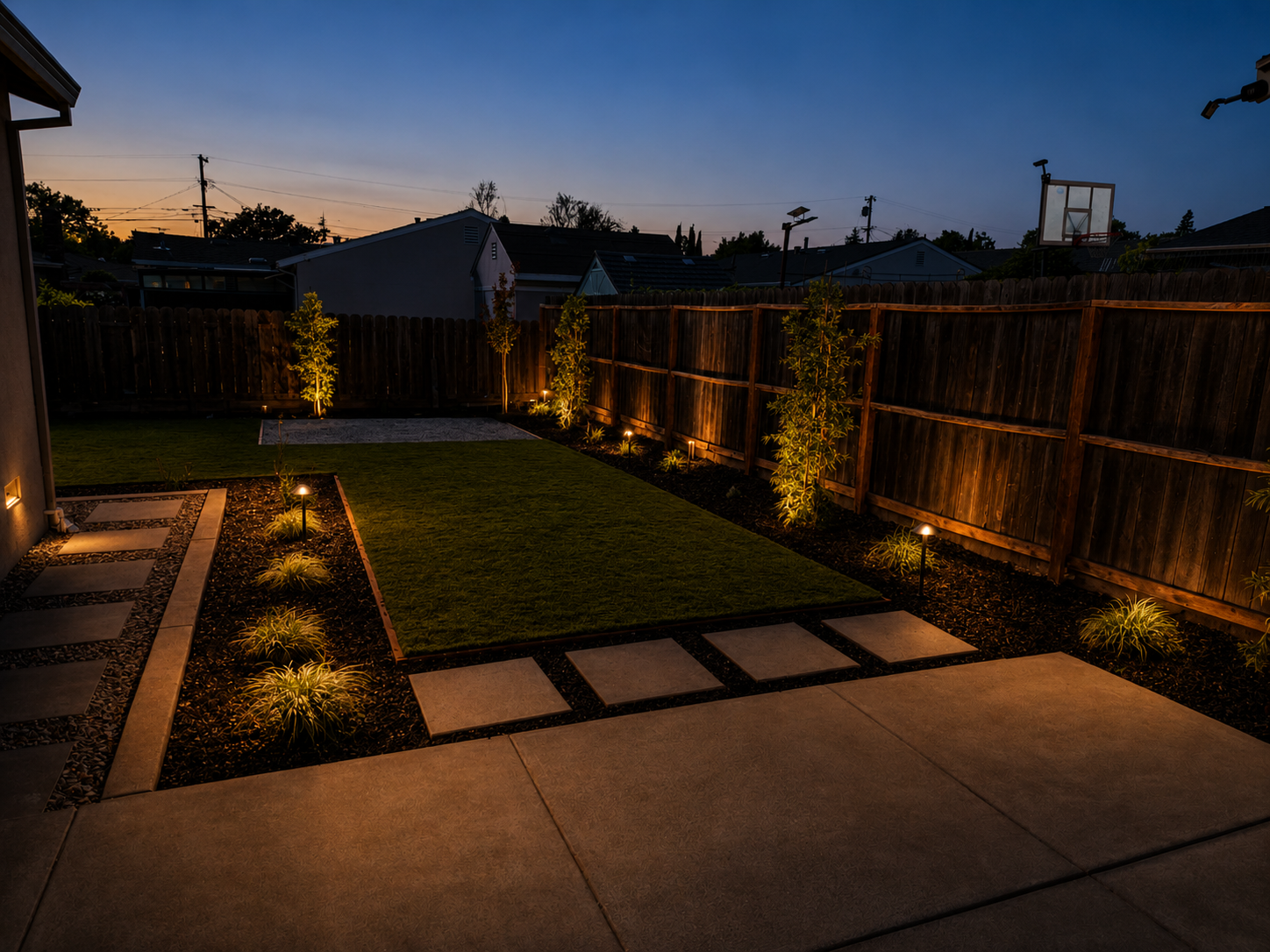 Nighttime view of a backyard with illuminated pathway lights, a small lawn, and a wooden fence. Tall trees are lit from below, and neighboring houses with rooftops and a basketball hoop are visible in the background. The sky is darkening with a hint 