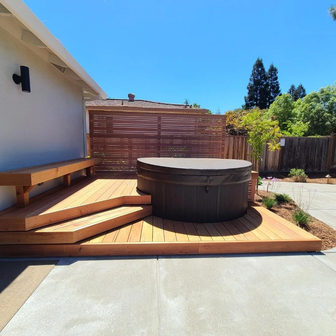 A backyard with a wooden deck, a round hot tub with a cover, a wooden bench along the house wall, and a privacy wooden fence. There are trees and plants in the background with a clear blue sky.