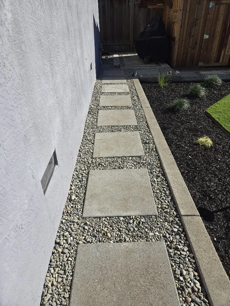 Concrete stepping stones on a gravel pathway alongside a white house wall and a garden with dark soil, plants, and a wooden fence.
