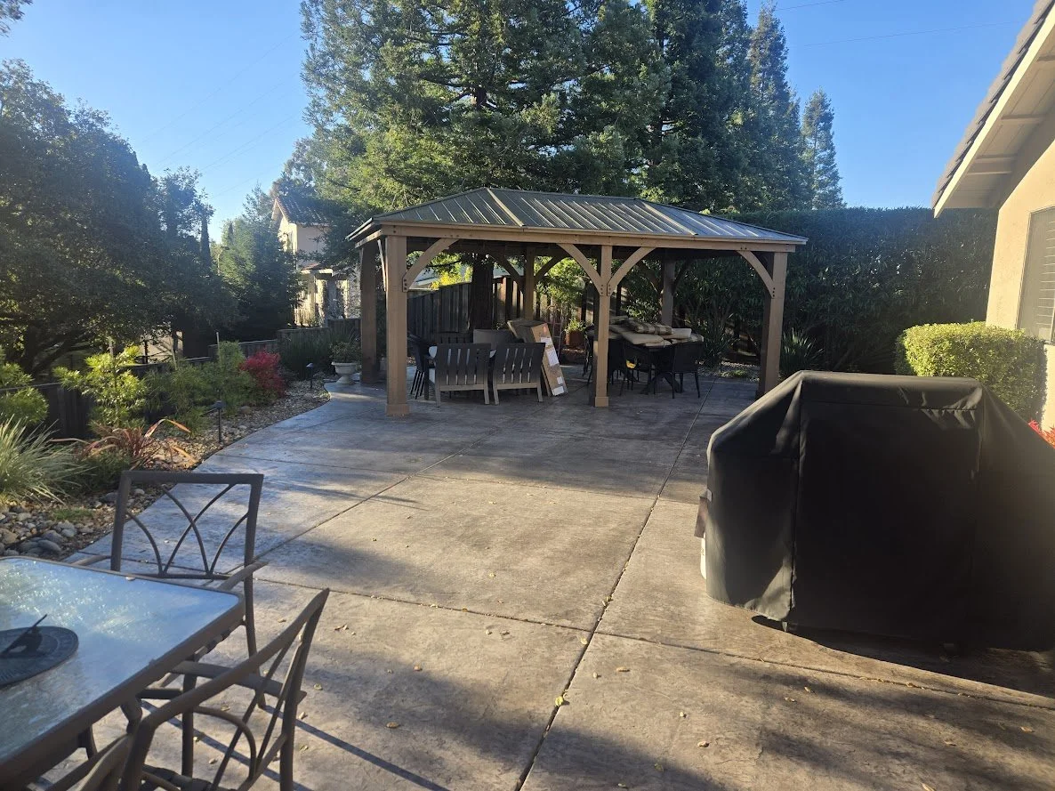 Backyard patio with a covered gazebo, outdoor furniture including a dining table and chairs, covered barbecue grill, surrounding plants, and a concrete patio surface under a clear blue sky.