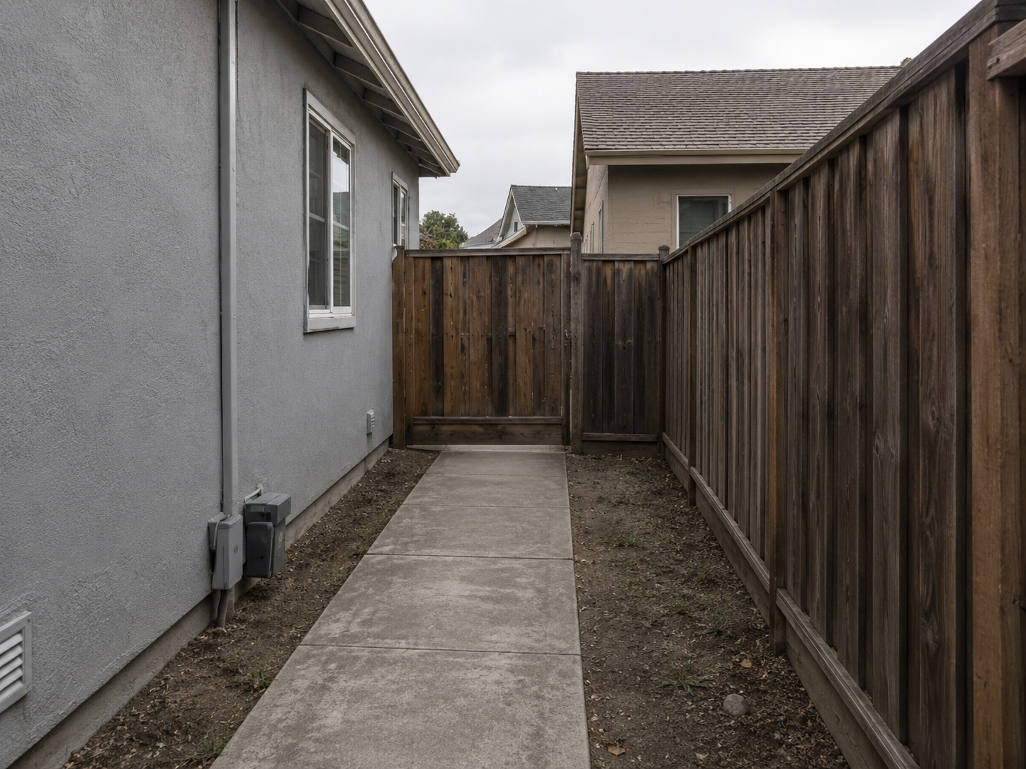 Side yard with a concrete walkway, grey house with windows on the left, and a tall wooden privacy fence on the right, leading to a wooden gate.
