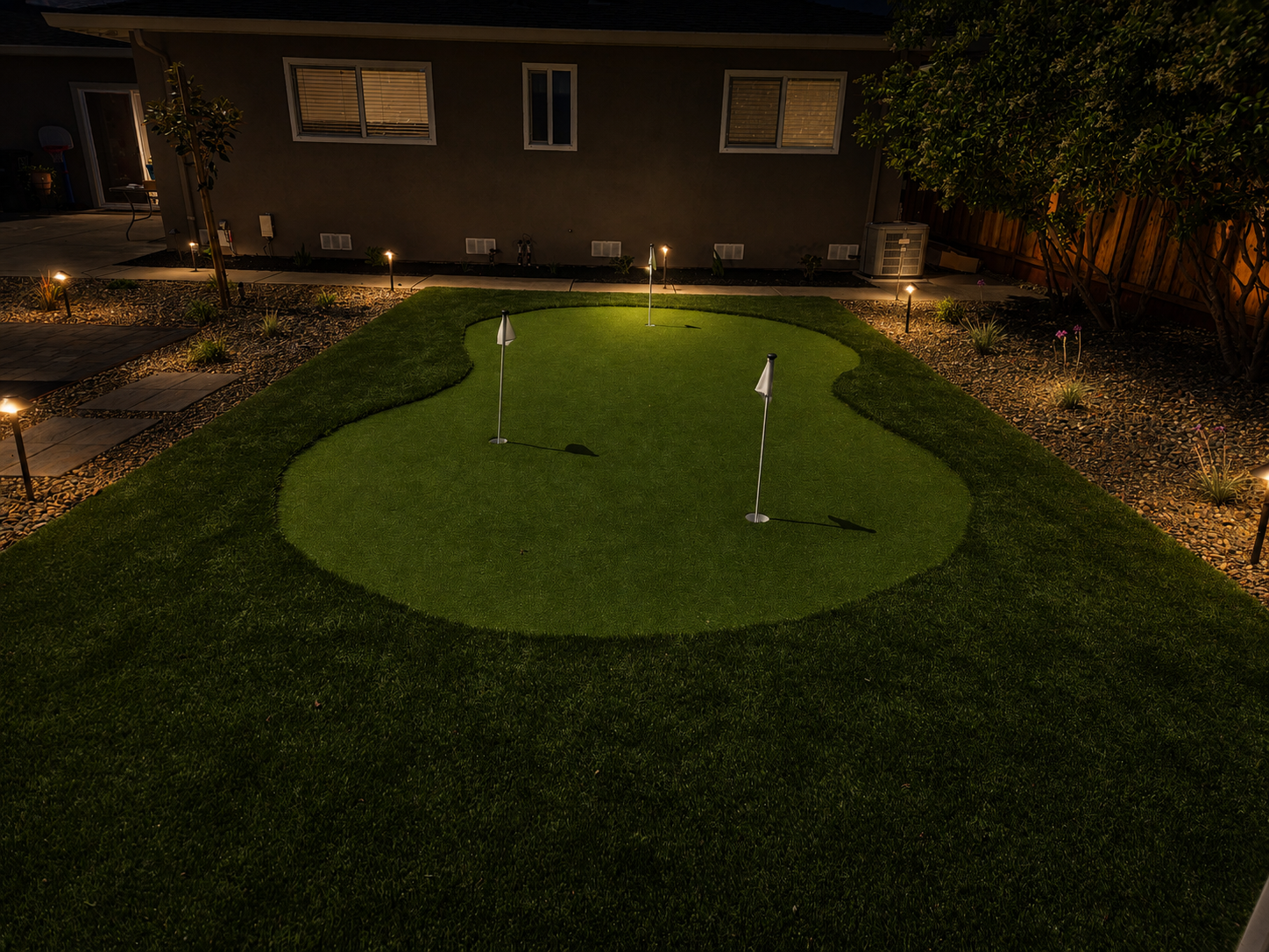 Nighttime backyard with a mini golf putting green, three small flags, and garden lights along the pathway