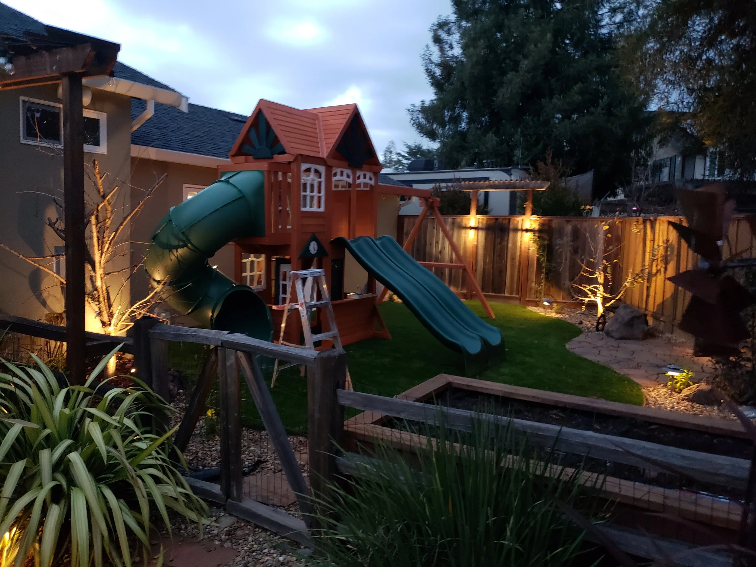 A backyard with a wooden playhouse and slide, surrounded by a fence, trees, and outdoor lighting at dusk.