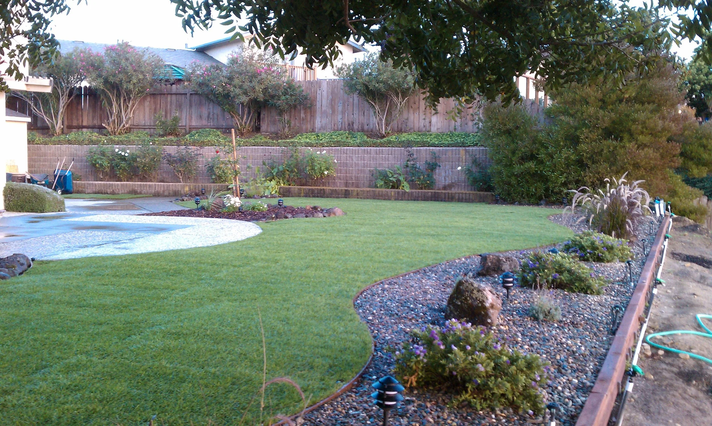 Residential backyard with a well-maintained lawn, flower beds, shrubs, trees, and a wooden fence.