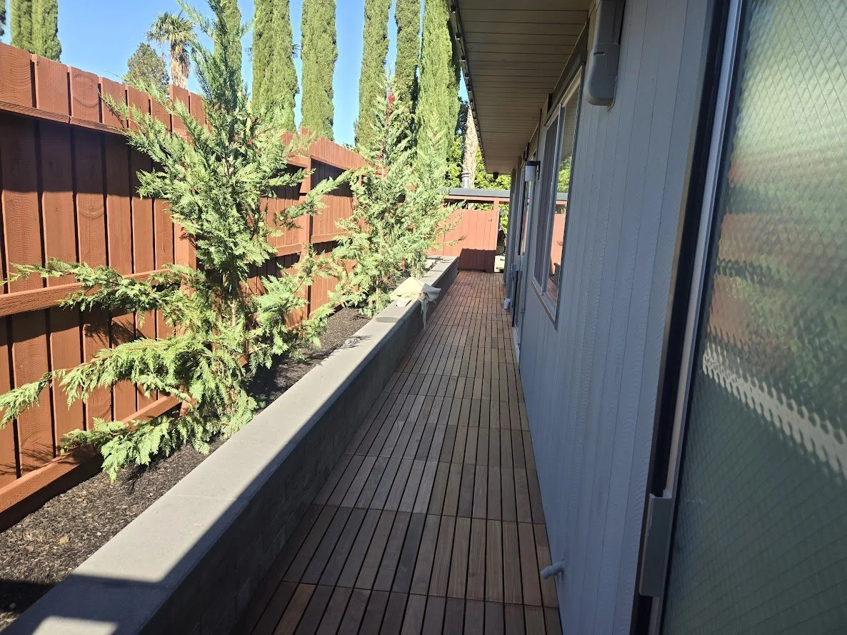 View of a wooden outdoor patio with a brown fence and potted evergreen trees along the side of a house, with a white exterior wall and windows, on a sunny day.