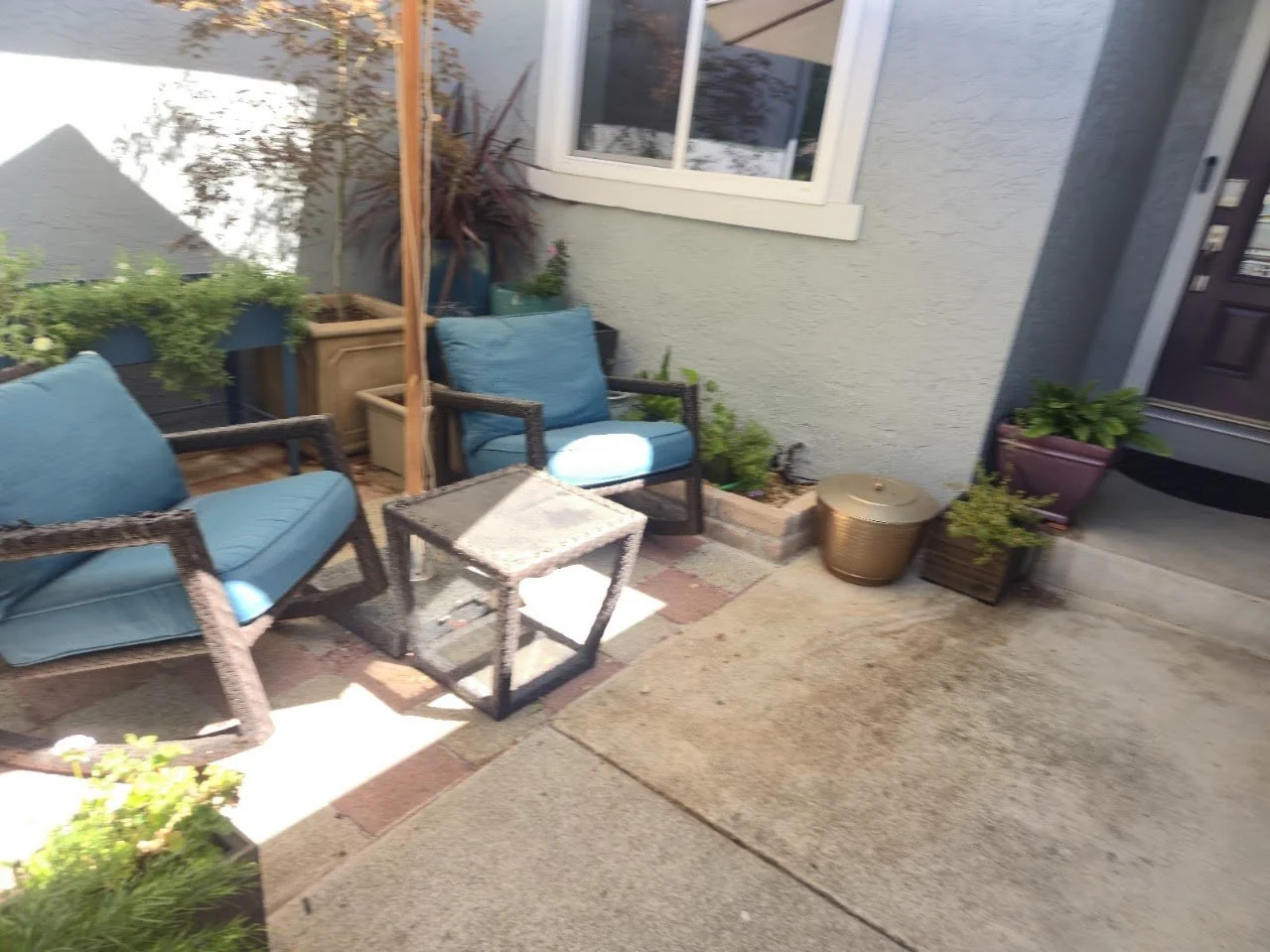 Outdoor patio area with two blue cushioned chairs, a small side table, and various potted plants near a house wall and window.