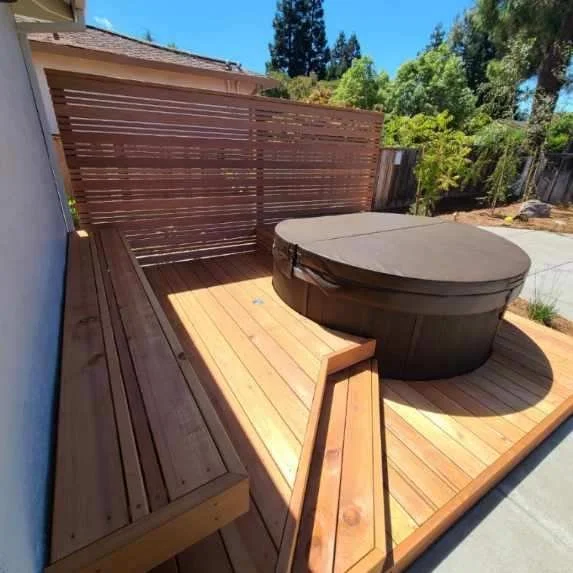 Wooden deck with a hot tub covered by a black cover, surrounded by wooden fencing and green trees in the background under a blue sky.