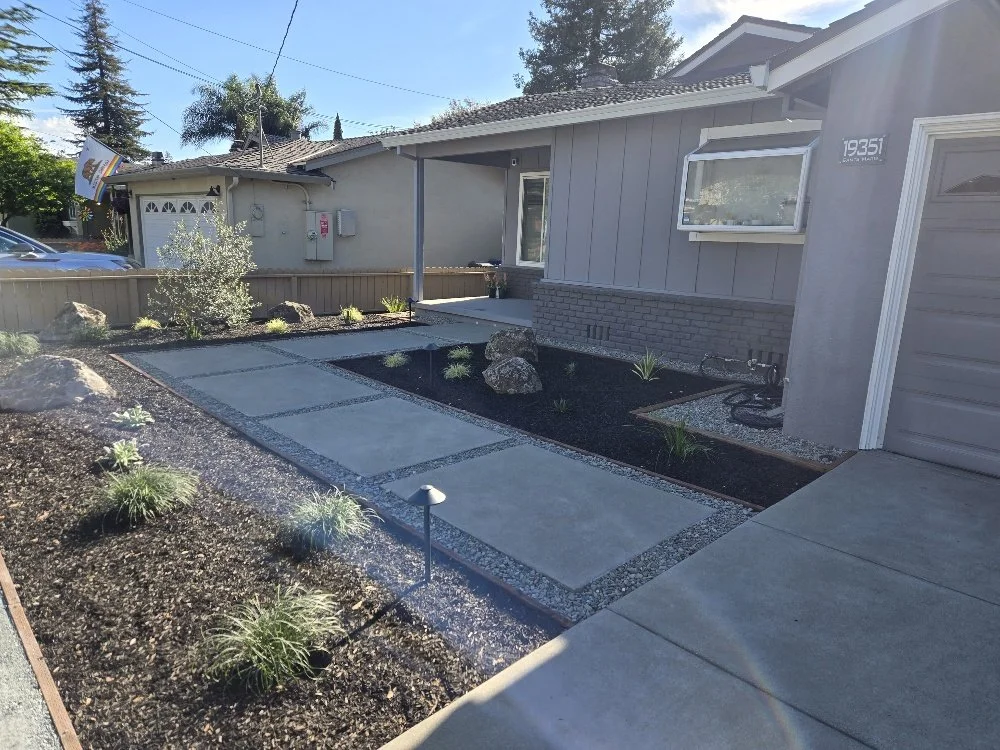 Front yard with newly paved concrete pathways, landscaping rocks, small plants, and a mulch bed, in front of a grey house with a garage door.