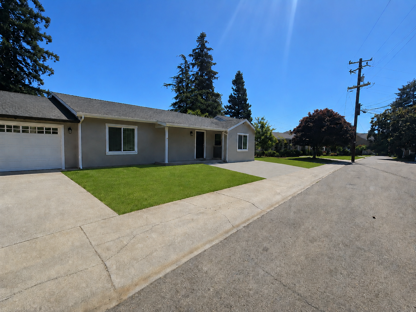 Single-story gray house with attached garage, front lawn with green grass, concrete driveway and sidewalk, trees in the background, power pole, clear blue sky, sunny day.
