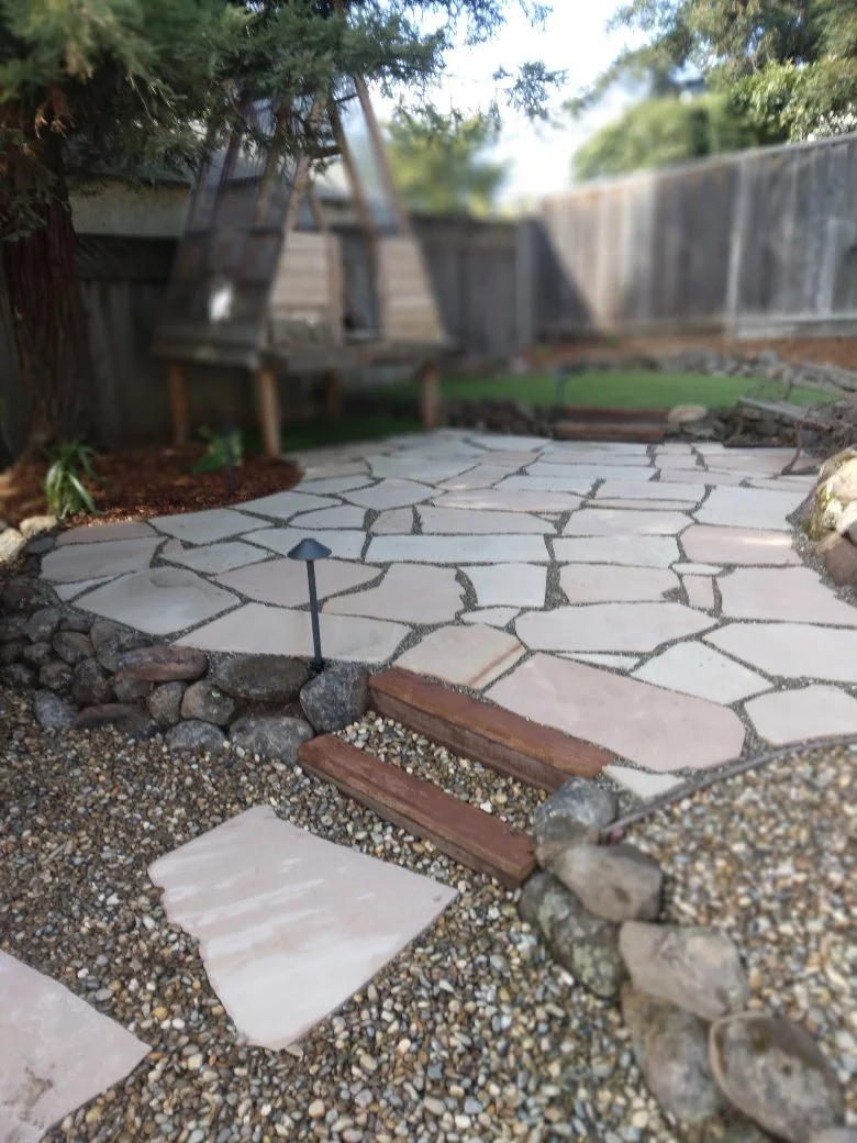 A backyard patio with irregularly shaped flagstone paving, gravel ground cover, and a wooden fence in the background. A small pathway with wooden steps and a solar-powered garden stake light are visible.