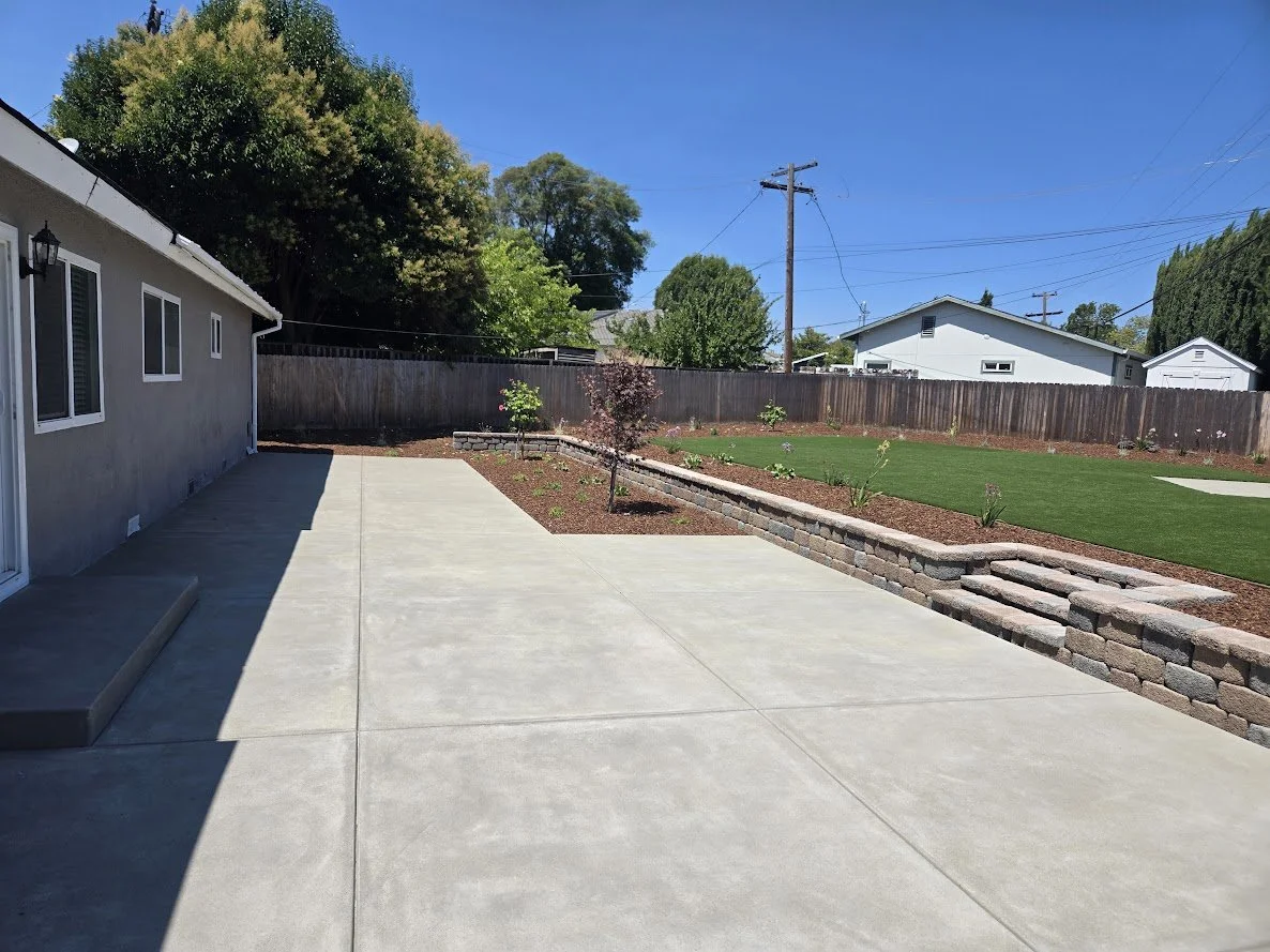 Backyard with concrete patio, a garden bed with small shrubs, and a lawn area bordered by a brick wall and a wooden fence, with houses and trees in the background under a clear blue sky.