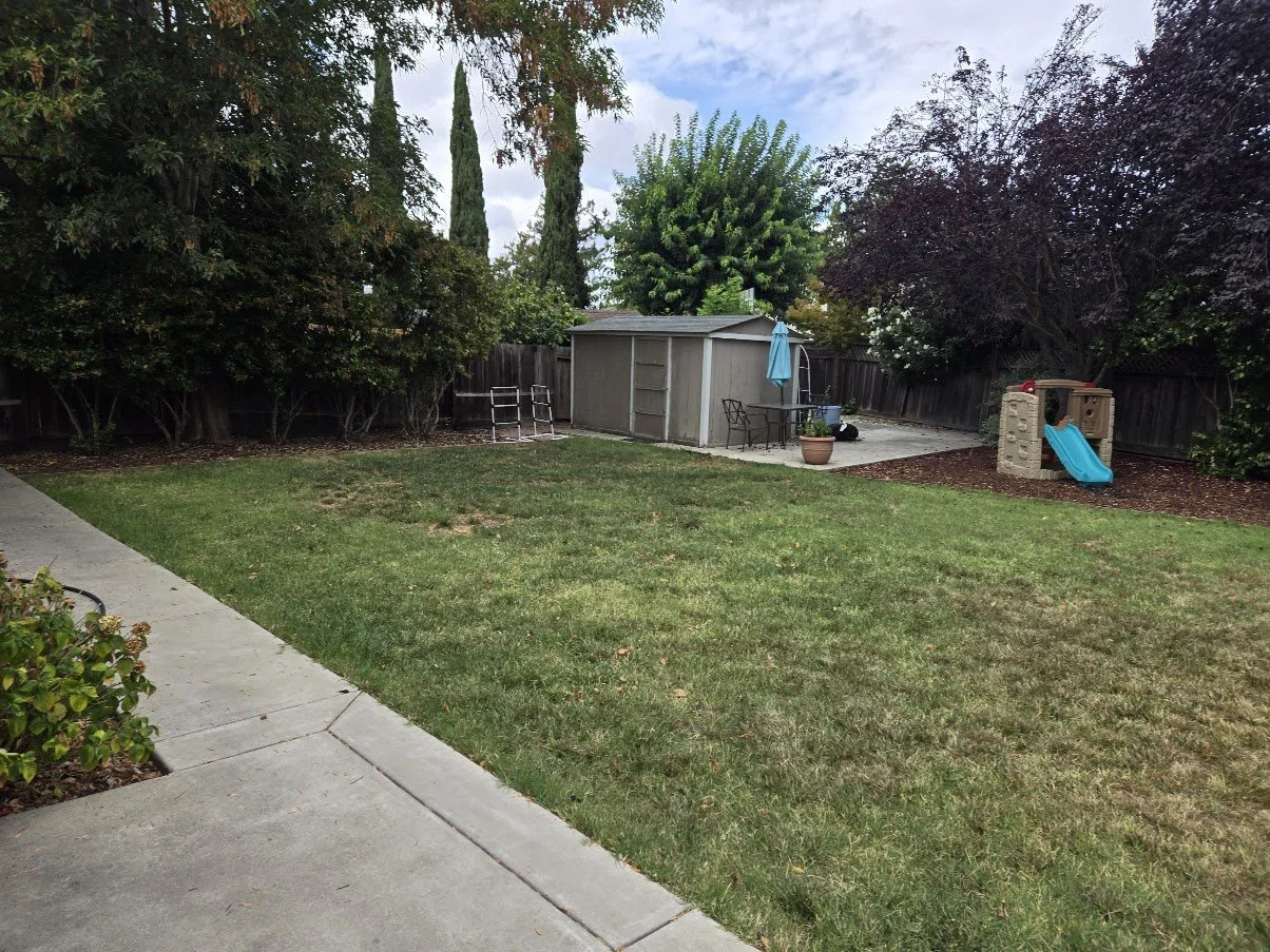 A backyard with a grassy lawn, trees, a shed, outdoor furniture with an umbrella, and a small plastic playhouse with a slide.