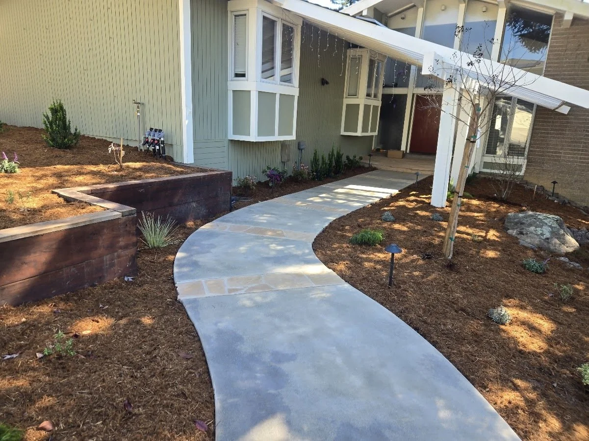 Curved concrete walkway leading to your house with a small landscaped yard on each side, some plants, small trees, and landscape lights, a wooden raised garden bed, and the house exterior with bay windows, a front door, and a porch.