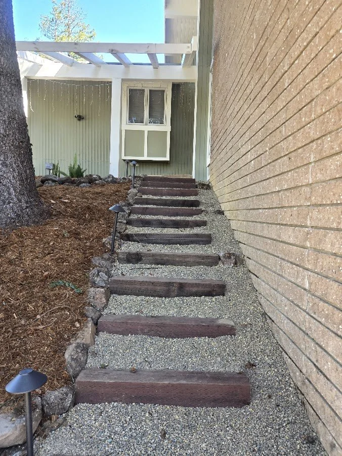 Outdoor gravel pathway with wooden steps leading up to a house, flanked by a brick wall on the right and a garden area on the left with large rocks and small solar lights.
