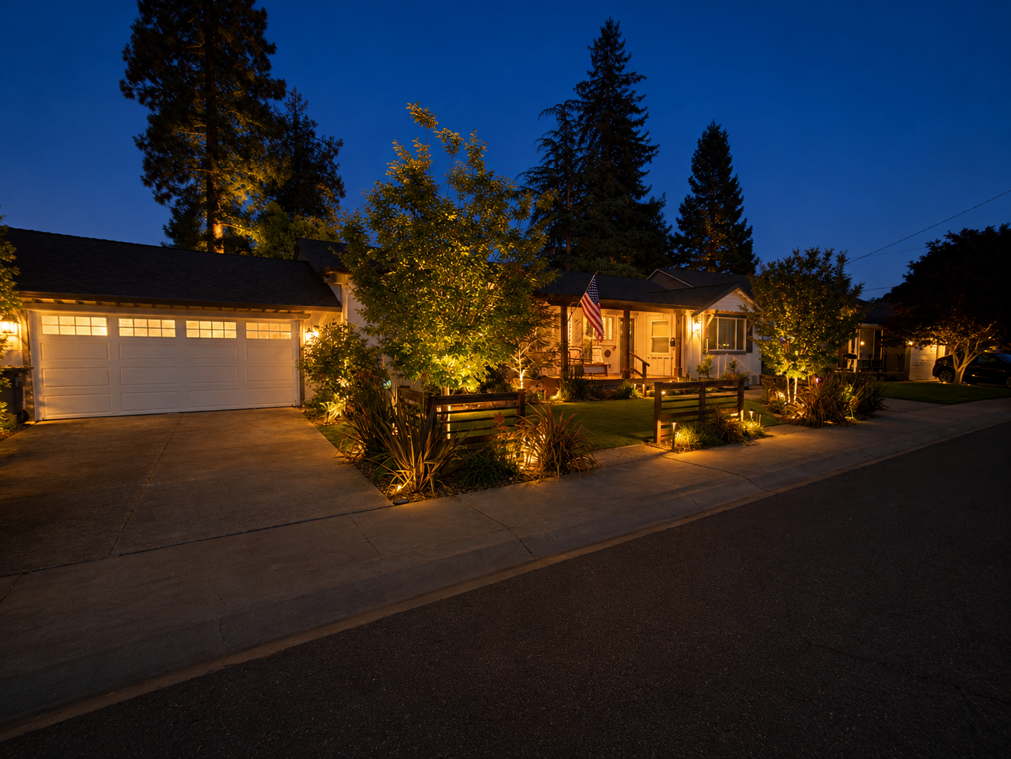 A cozy suburban house at night with warm exterior lighting, a front porch, an American flag, and landscaped yard with trees and bushes.